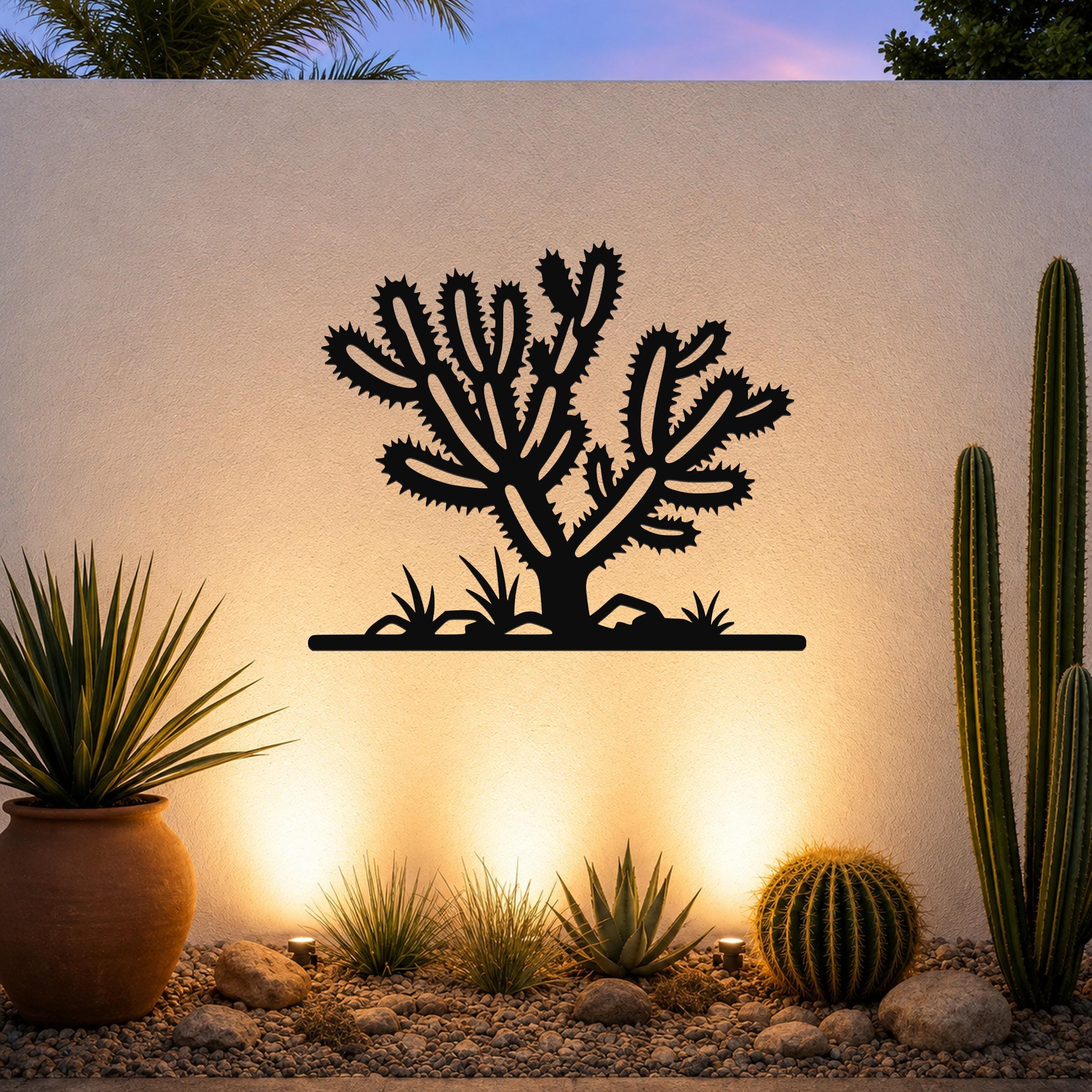 Black metal Cholla cactus wall art with spiky branch cutouts, mounted on an outdoor wall above a desert garden with potted plants and ground lights.
