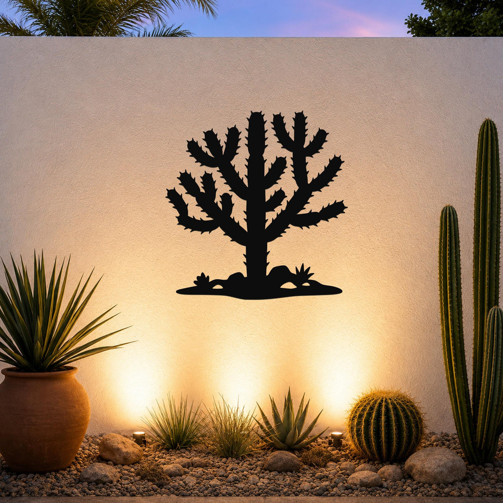 Black metal Cholla cactus wall art silhouette with spiny branches and ground cutout, mounted on a textured outdoor wall above a desert garden with potted plants and ground lights.