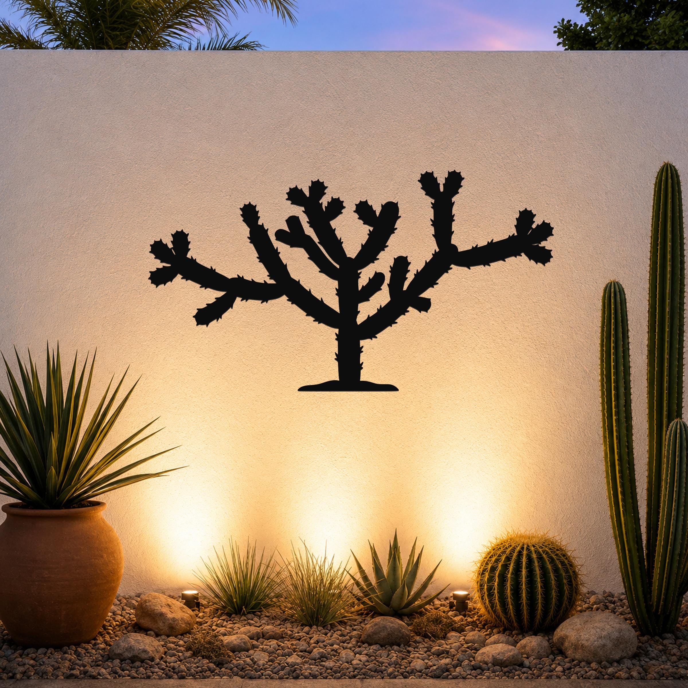 Black metal Cholla cactus wall art silhouette with branching arms and spines, mounted on a textured outdoor wall above a desert garden with potted plants and ground lights.