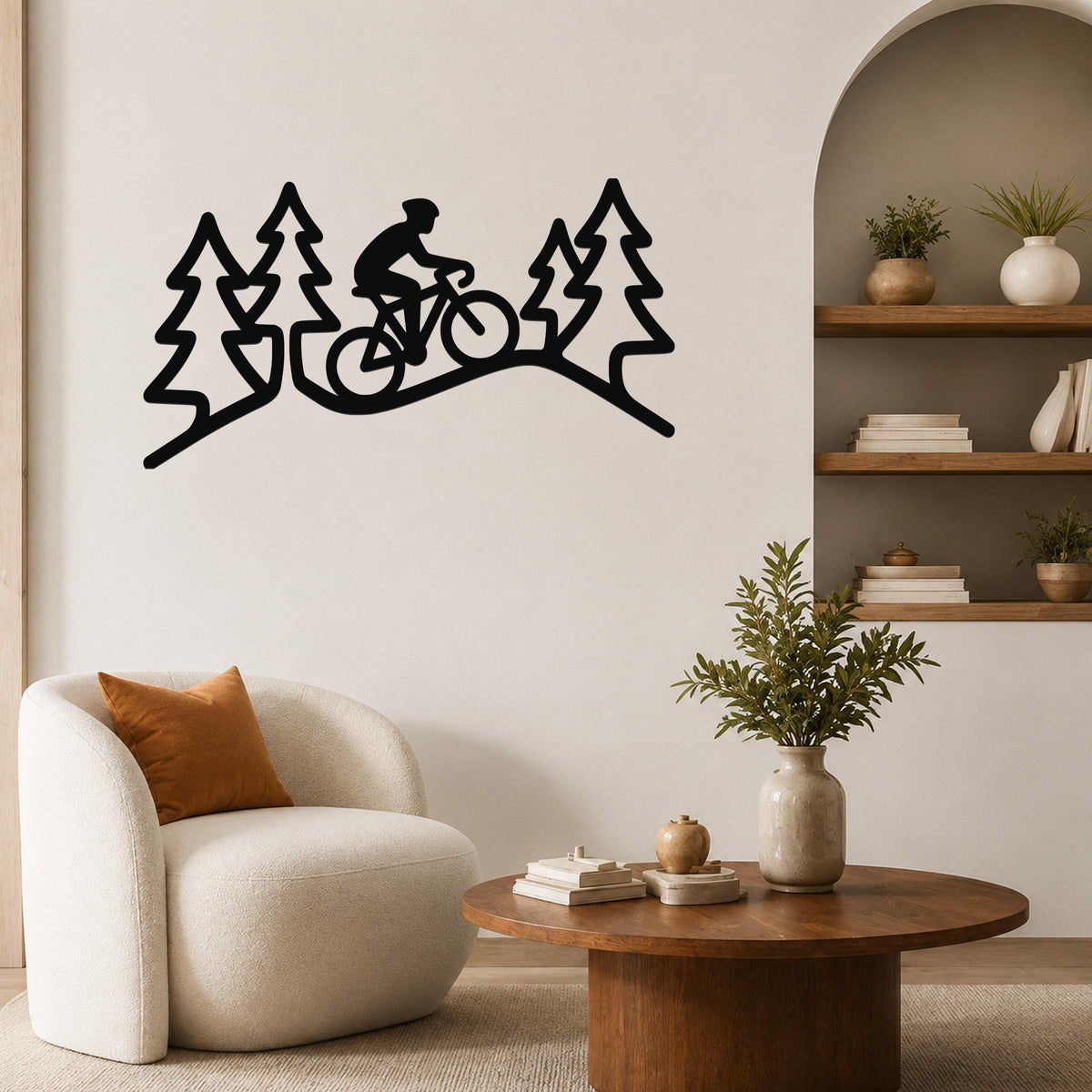 Black metal wall art featuring a cyclist silhouette riding through a forest of pine trees, mounted on a living room wall above a white armchair.