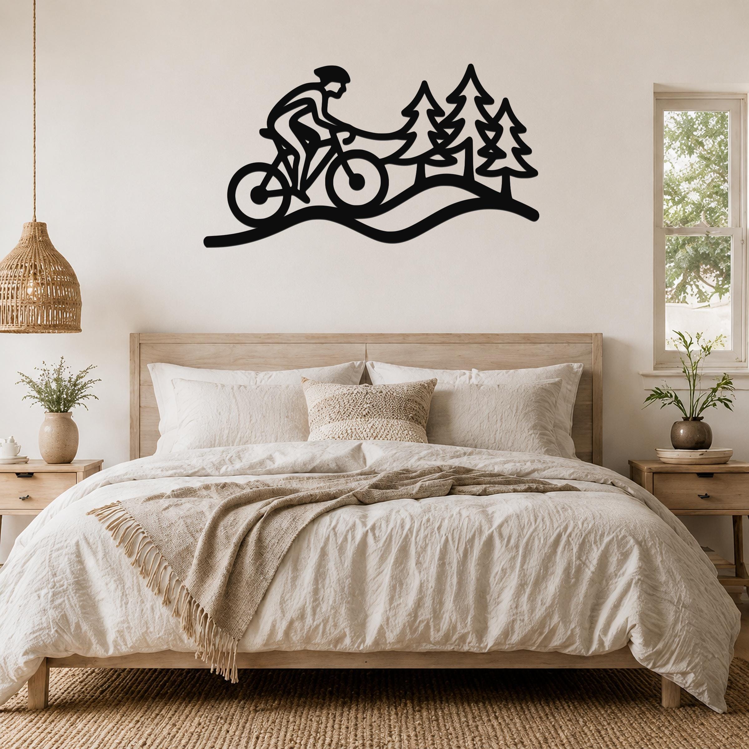 Black metal wall art silhouette of a cyclist on a trail with pine trees, mounted above a bed in a bedroom.