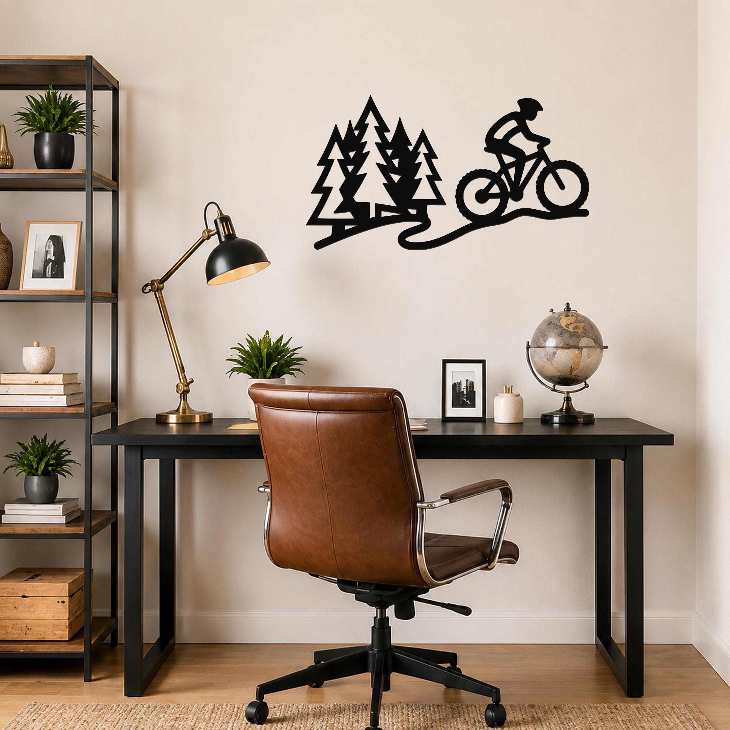 Black metal wall art cutout of a cyclist on a trail with pine trees, mounted above a black desk in a home office.
