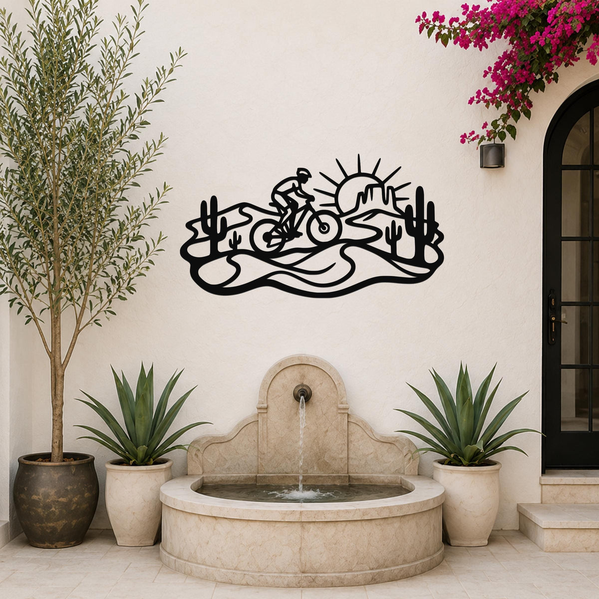 Black metal wall art silhouette of a cyclist on a desert trail with cacti and a sun, mounted on a white outdoor wall above a stone fountain.
