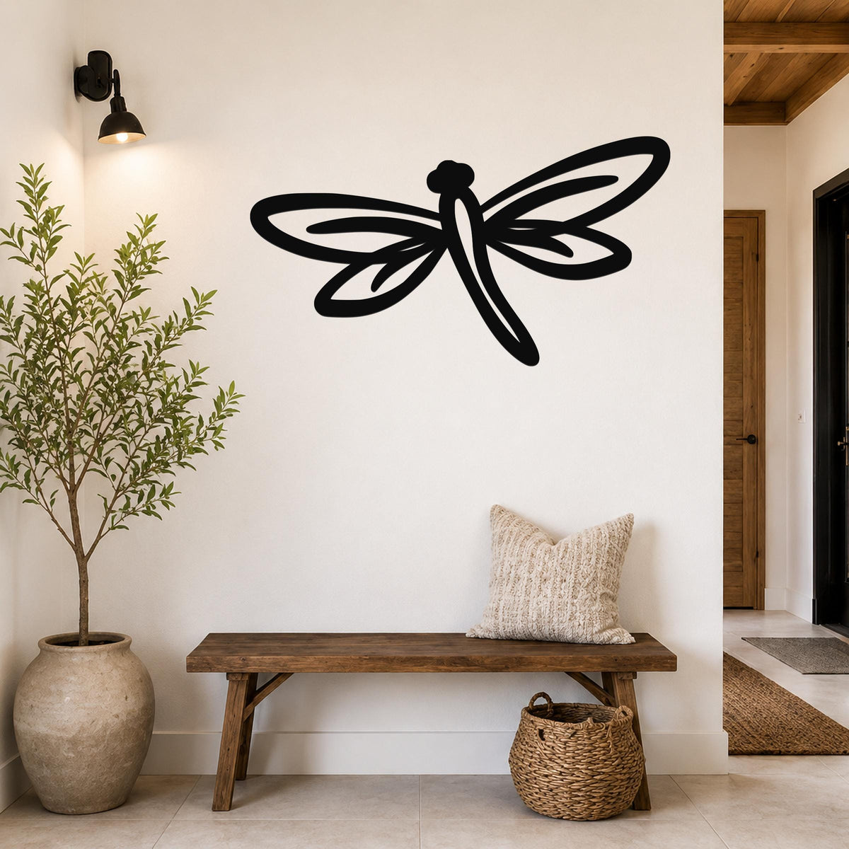 Black metal dragonfly wall art with cutout wings and body, mounted on a white wall above a wooden bench in a hallway.