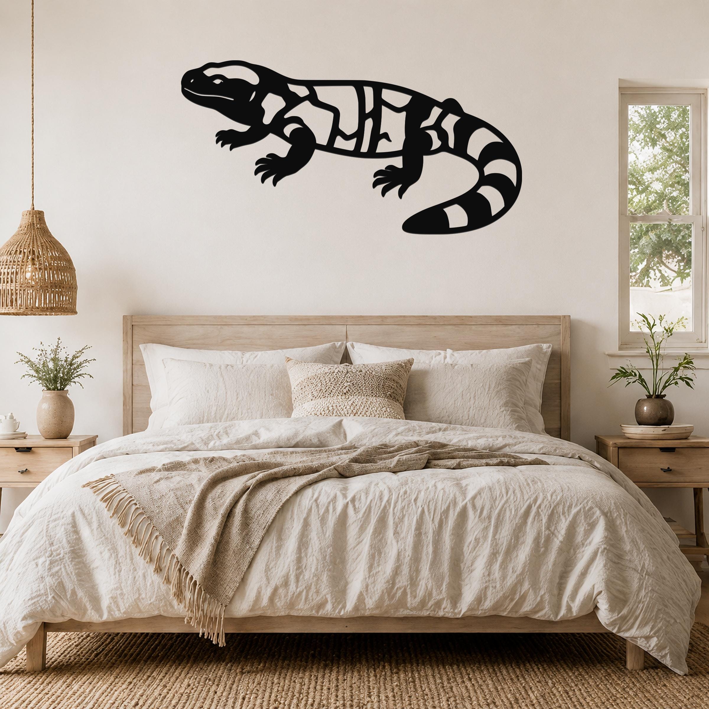 Black metal Gila monster wall art with internal cutout patterns, mounted above a bed in a neutral bedroom.