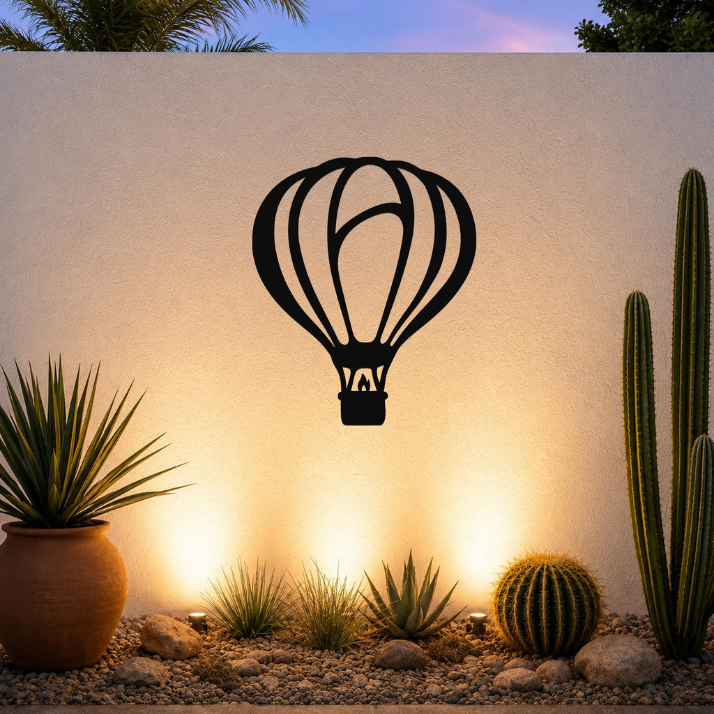 Black metal hot air balloon wall art with internal flame cutout, mounted on an outdoor wall above a desert garden with cacti and warm uplighting.