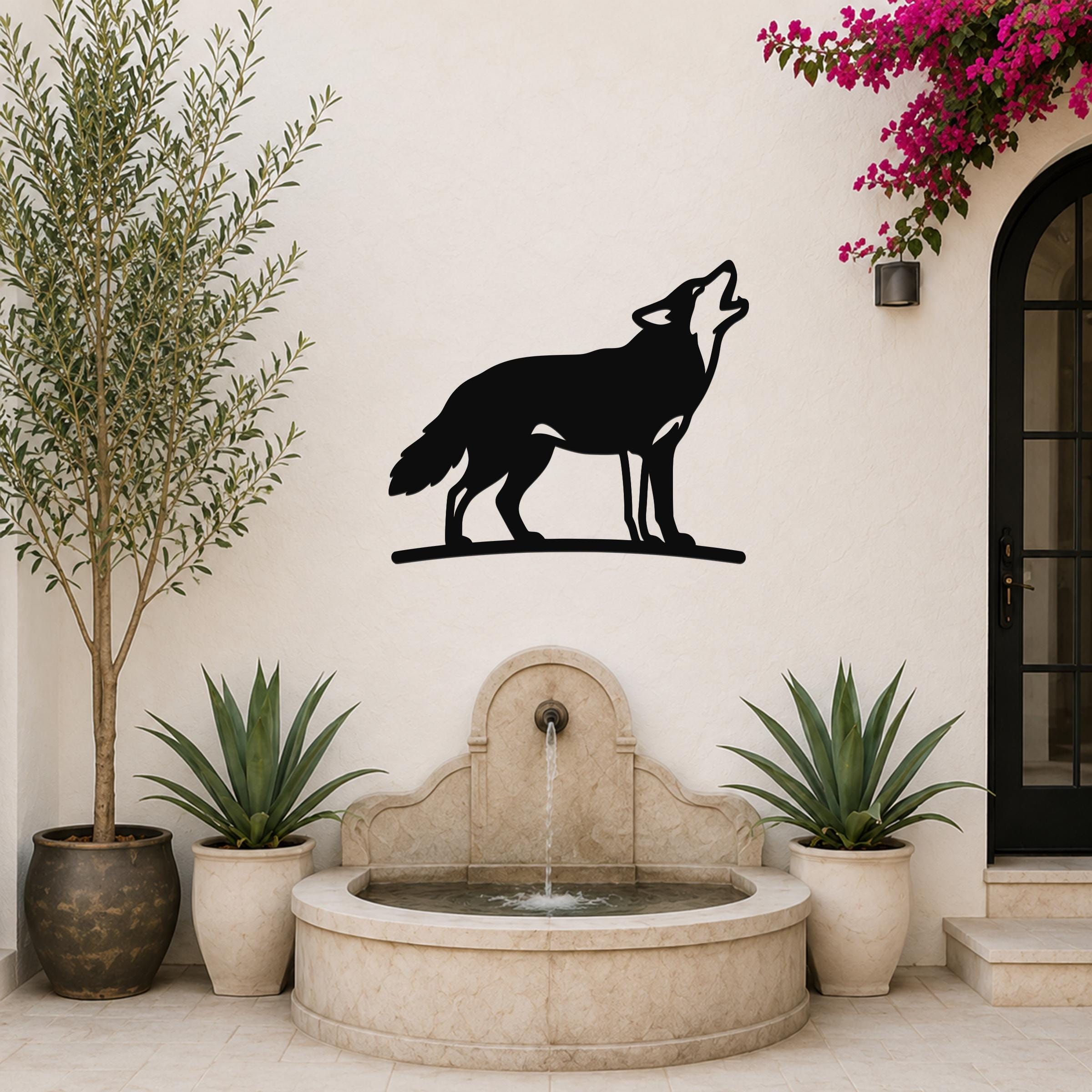 Black metal coyote wall art in a howling silhouette, mounted on a white outdoor wall above a stone fountain with potted plants and a flowering vine.