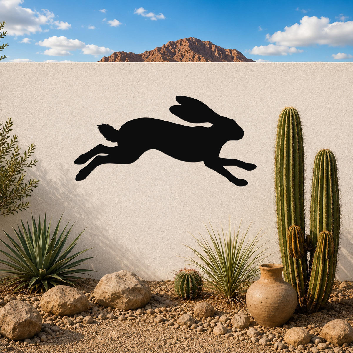Black metal jackrabbit wall art silhouette in a leaping pose, mounted on a white outdoor wall above a desert garden with cacti and rocks.