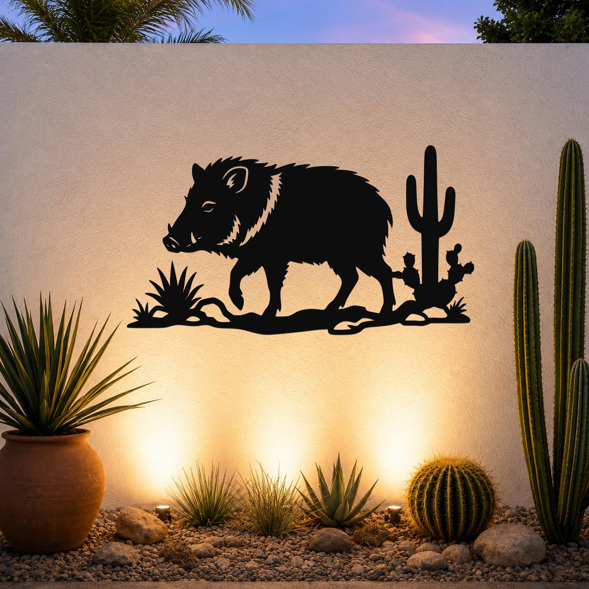 Black metal wall art silhouette of a javelina with cacti, mounted on an outdoor wall above a desert garden with potted plants and ground lights.