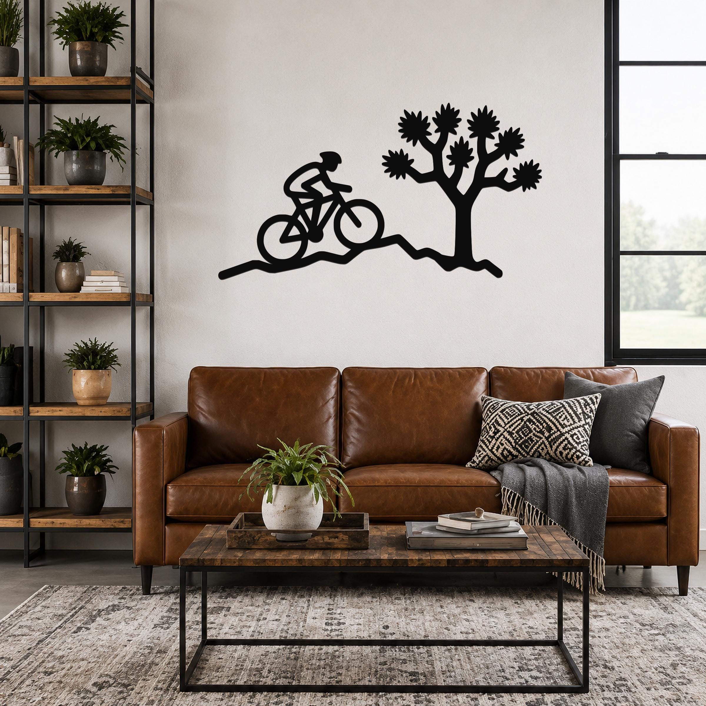 Black metal wall art silhouette of a cyclist on a trail beside a Joshua tree, mounted above a brown leather sofa in a living room.