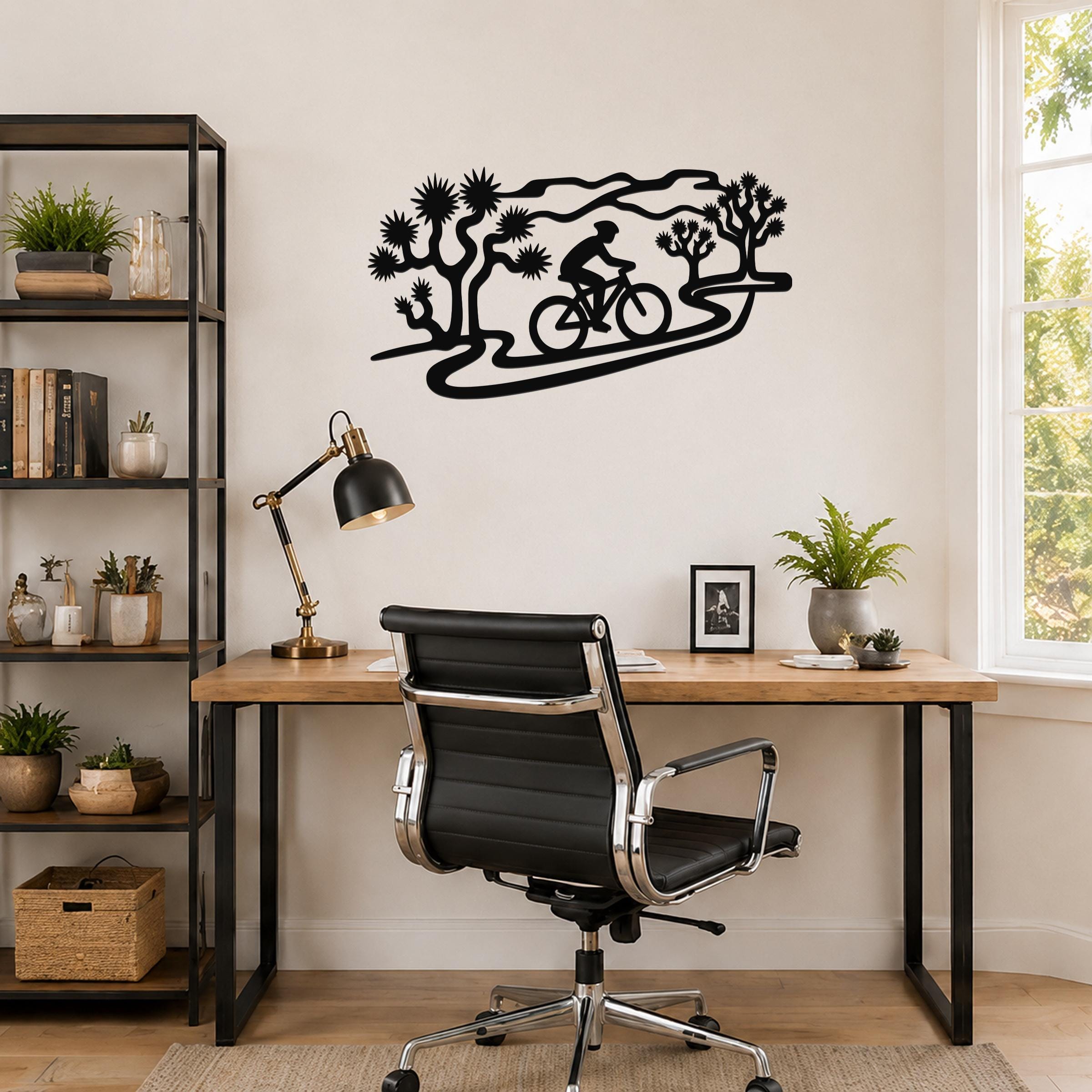 Black metal wall art silhouette of a cyclist on a trail surrounded by Joshua trees, mounted on a wall above a wooden desk in a home office.
