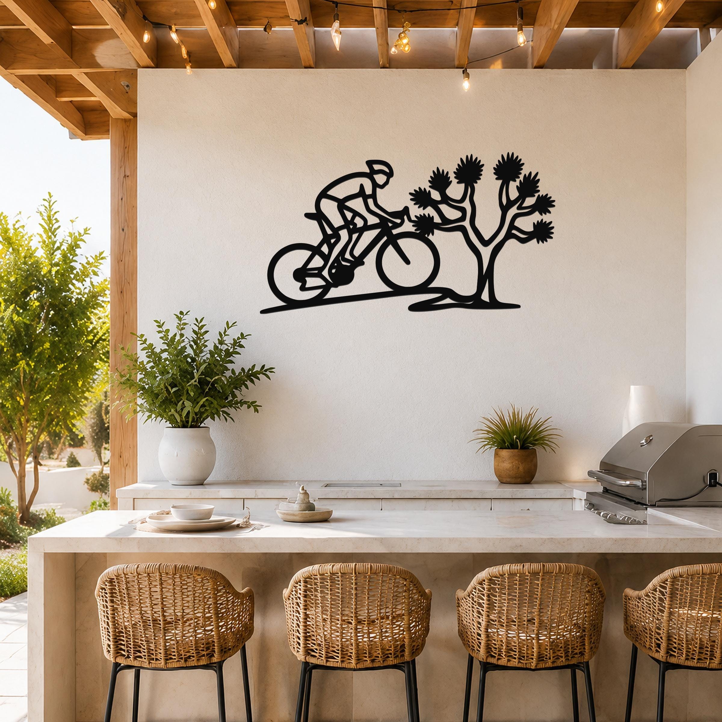 Black metal wall art cutout of a cyclist riding past a Joshua tree, mounted on a white wall above a kitchen counter with wicker bar stools.