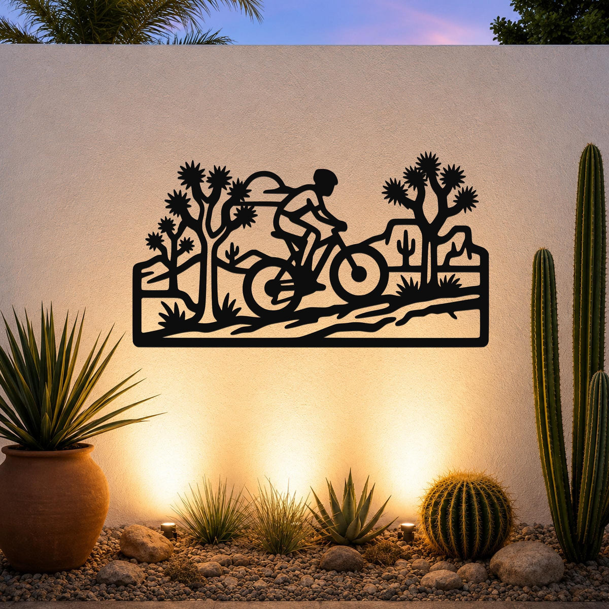 Black metal wall art cutout of a cyclist on a desert trail with Joshua trees and cacti, mounted on an outdoor wall above a gravel garden with potted succulents.