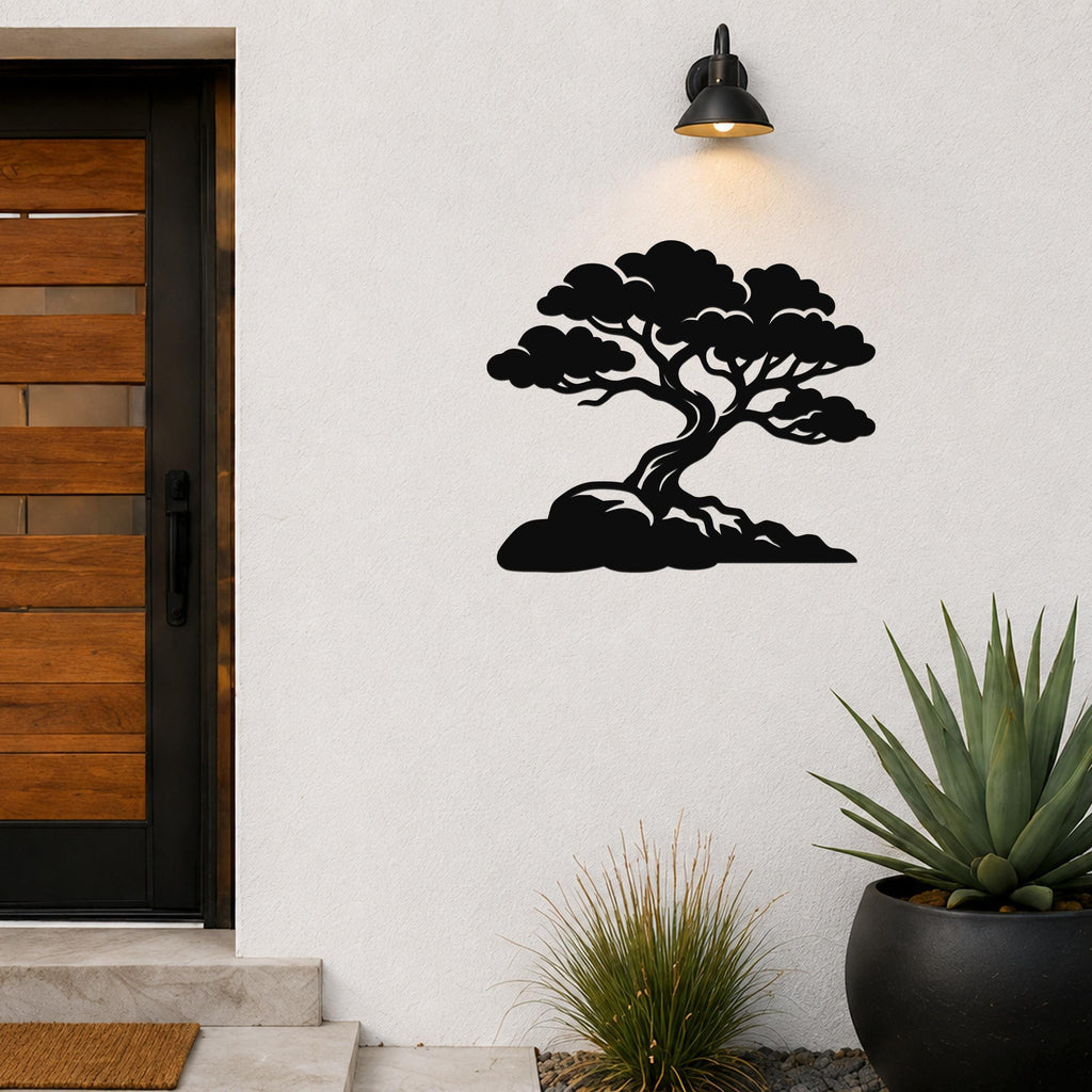 Black metal wall art cutout of a bonsai-style juniper tree with layered branches and roots, mounted on a white exterior wall above a potted succulent and near a wooden door.