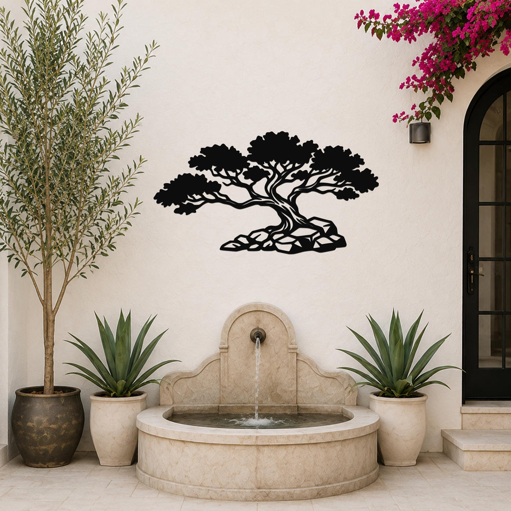 Black metal wall art in a bonsai tree silhouette with rocky base, mounted on a light-colored outdoor wall above a stone fountain.