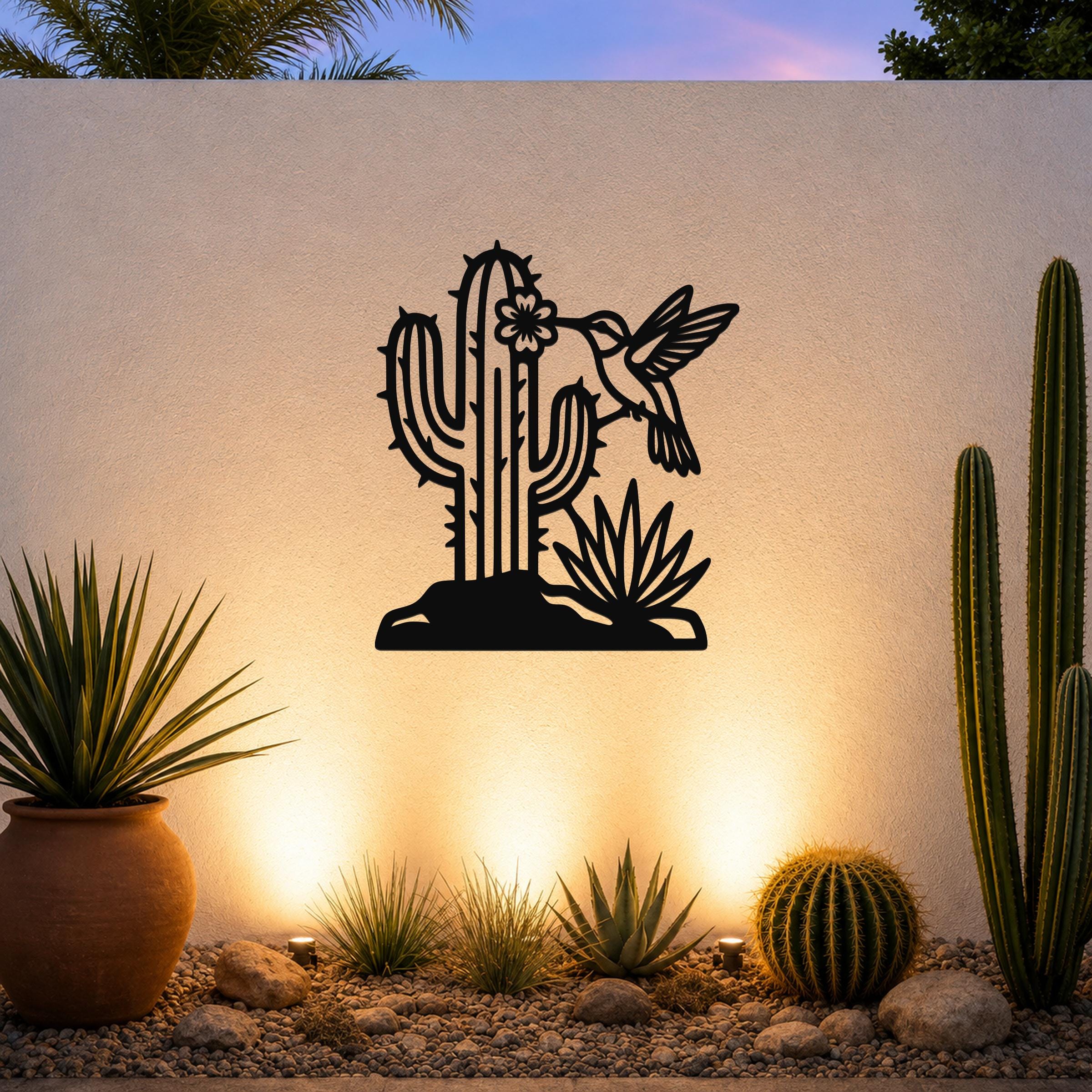 Black metal wall art with a cactus, flower, and hummingbird cutout design, mounted on a white outdoor wall above a desert garden with potted plants and ground lights.