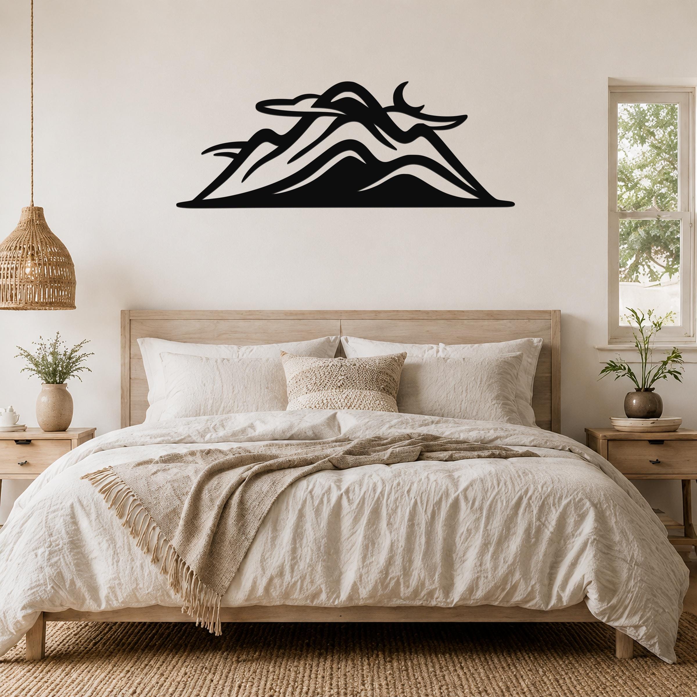 Black metal mountain silhouette wall art with layered peaks and a crescent moon cutout, mounted above a wooden bed in a neutral bedroom.