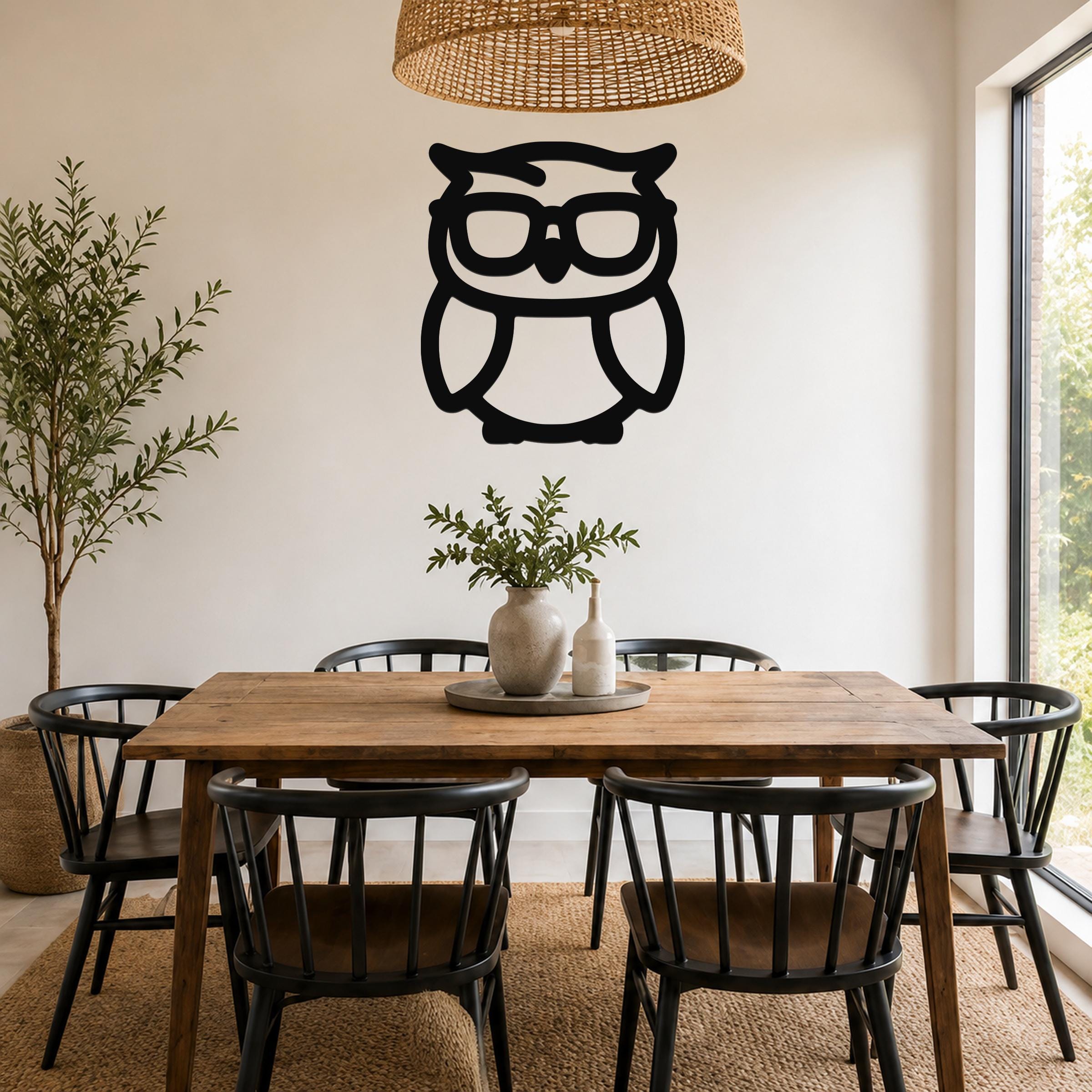 Black metal owl wall art with glasses silhouette, mounted on a wall above a wooden dining table in a bright room.