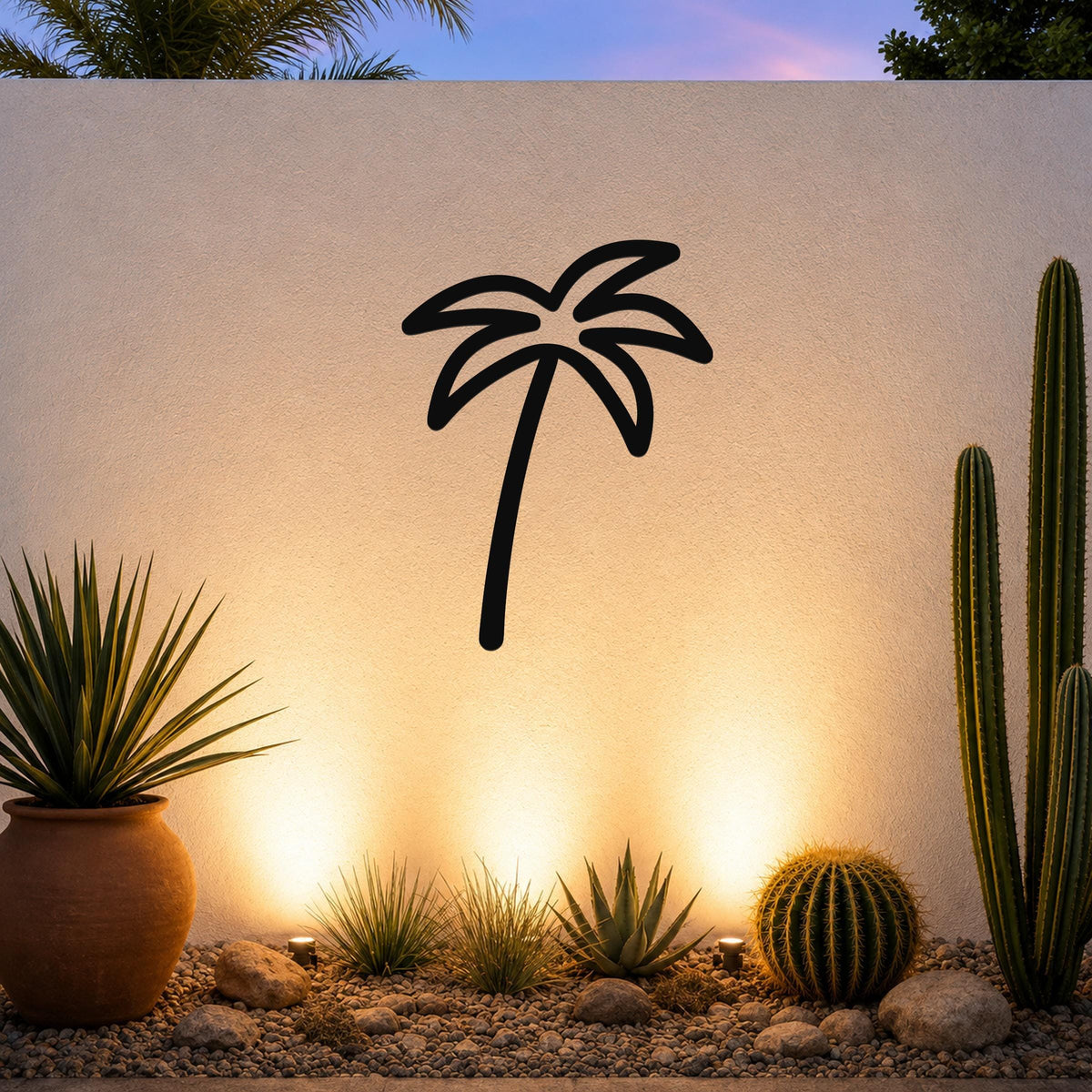 Black metal palm tree wall art silhouette mounted on a white outdoor wall above a desert garden with cacti and warm uplighting.
