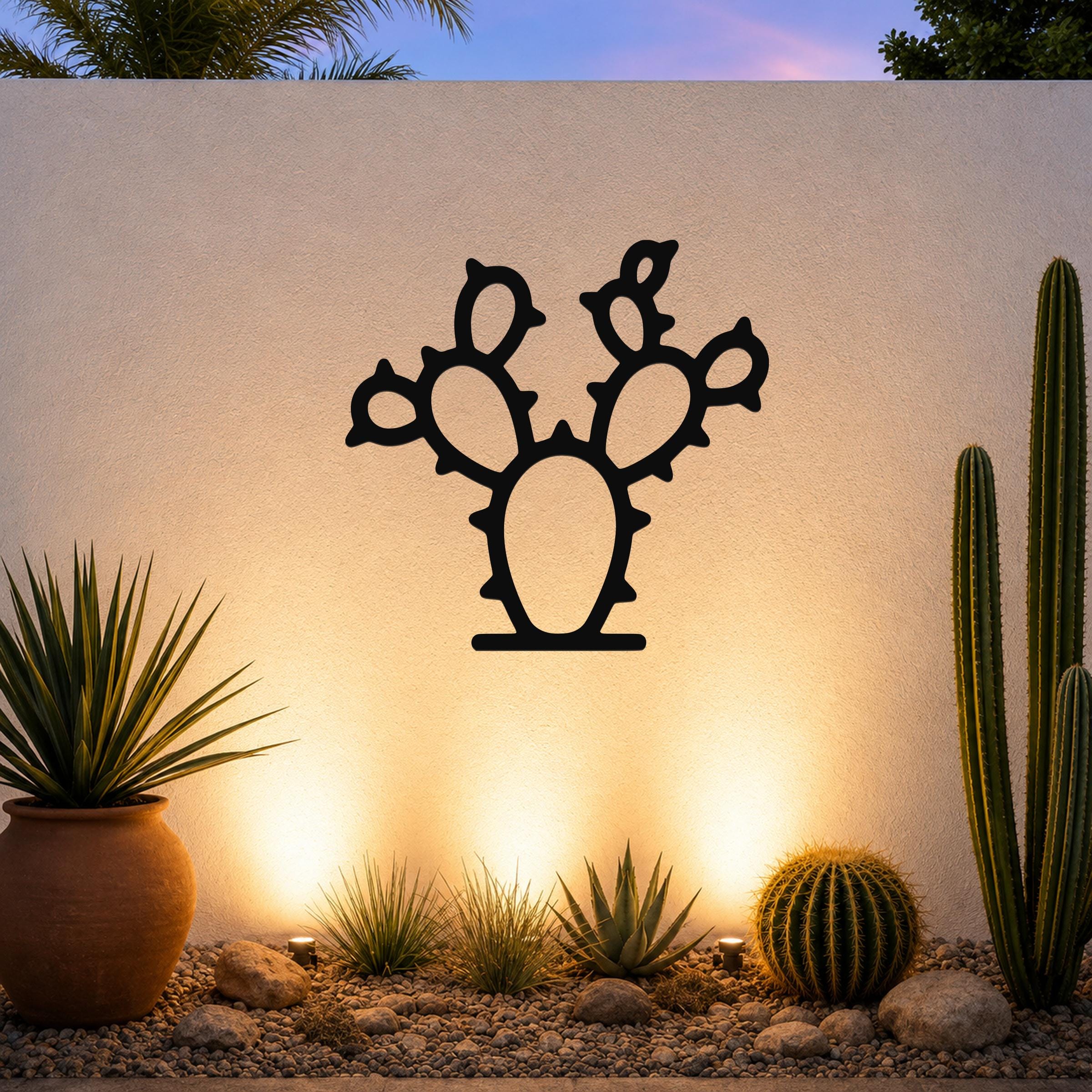 Black metal prickly pear cactus wall art with cutout pads and spines, mounted on a textured outdoor wall above a desert garden with potted plants and ground lights.