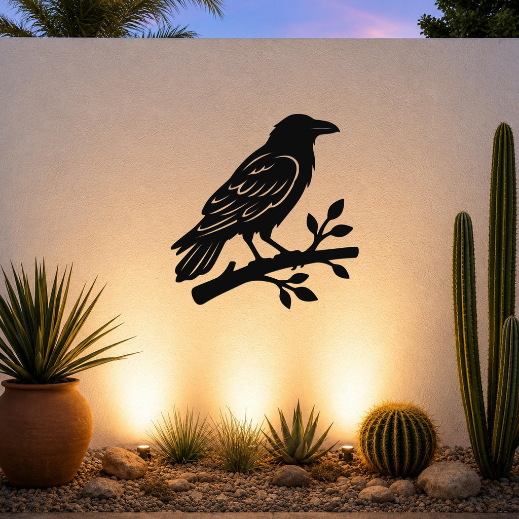 Black metal raven wall art silhouette perched on a branch, mounted on an outdoor wall above a desert garden with cacti and warm uplighting.