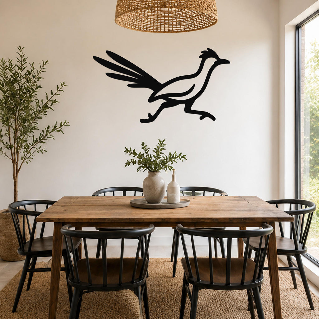 Black metal roadrunner wall art silhouette with spread wings, mounted on a white wall above a wooden dining table in a bright dining room.