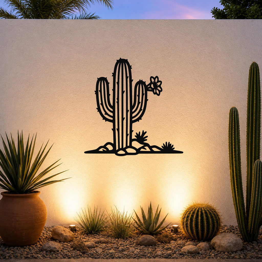 Black metal wall art silhouette of a saguaro cactus with flower and smaller cacti, mounted on an outdoor wall above a gravel garden with potted plants and uplighting.