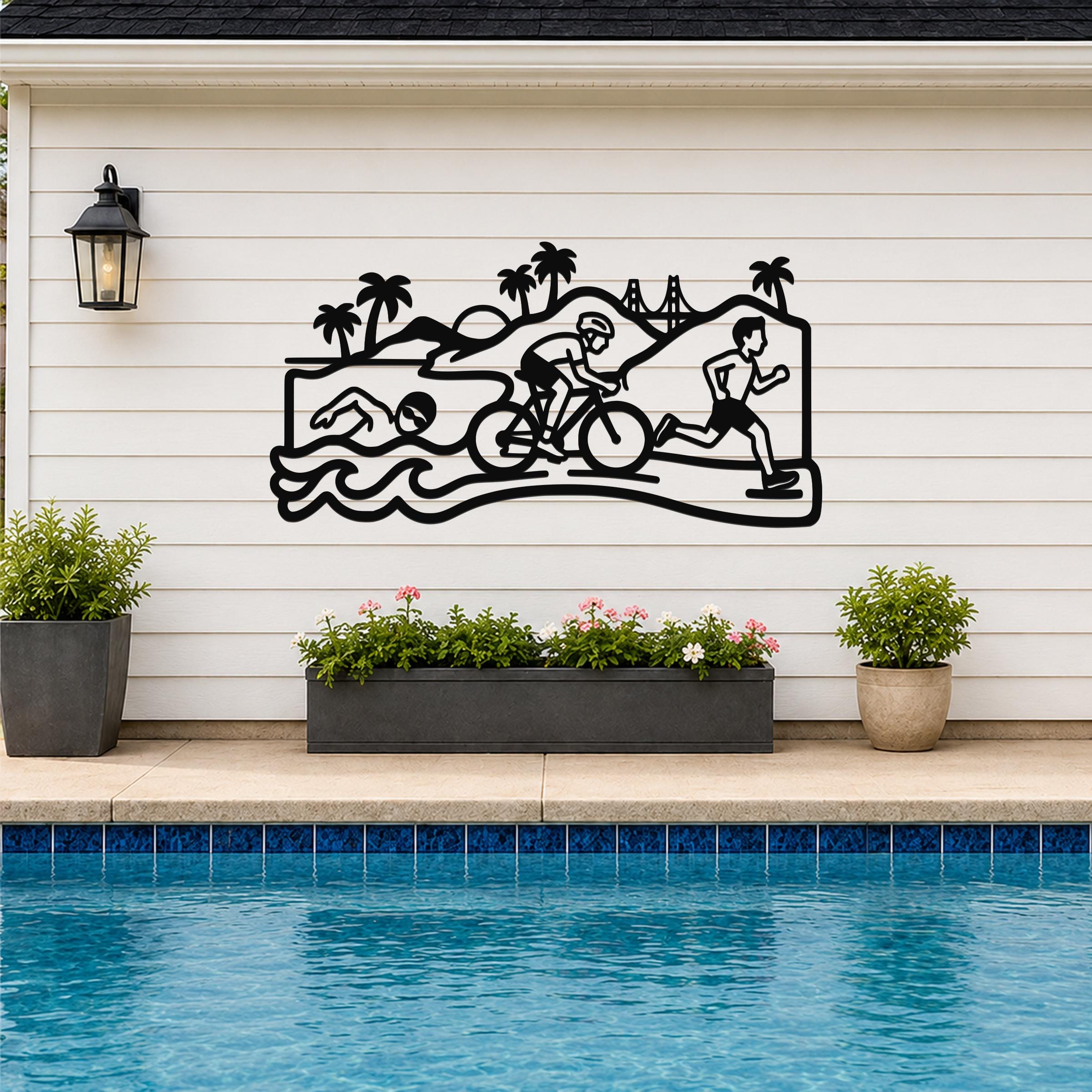 Black metal wall art depicting a triathlon scene with swim, bike, and run silhouettes, mounted on a white siding wall above a pool with potted plants.