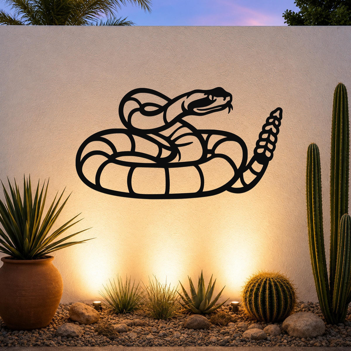 Black metal rattlesnake wall art with coiled silhouette and detailed tail rattle, mounted on an outdoor wall above a desert garden with cacti and warm uplighting.