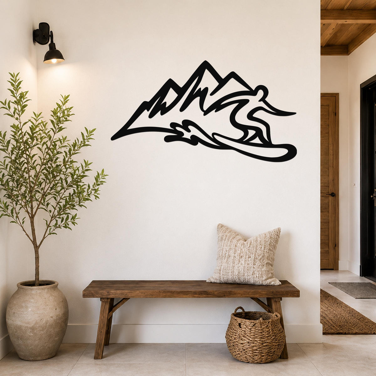 Black metal wall sculpture of a snowboarder in motion above a mountain range, mounted on a white wall above a wooden bench in a hallway.