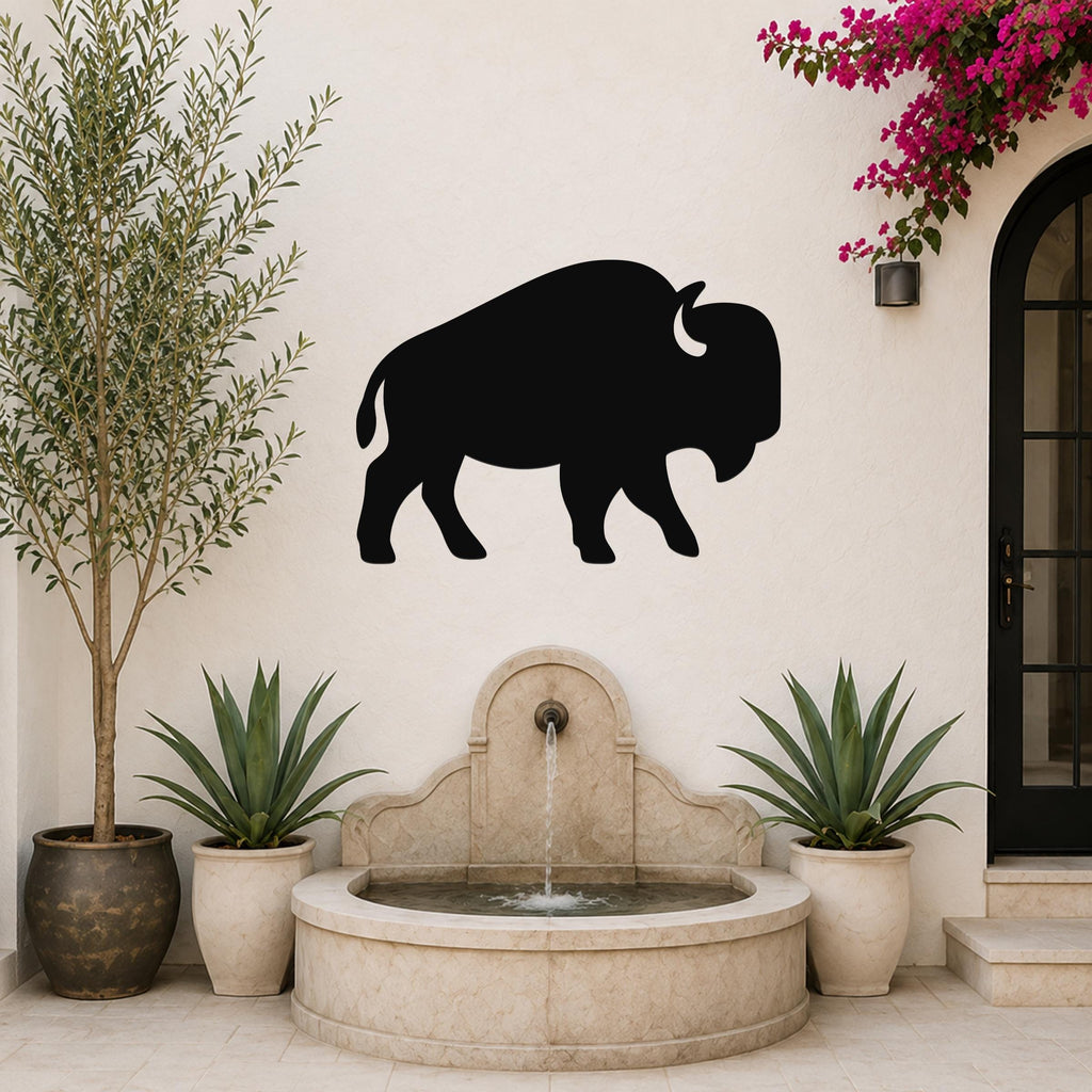 Black metal buffalo silhouette wall art mounted on a white outdoor wall above a stone fountain, flanked by potted plants and a flowering vine.