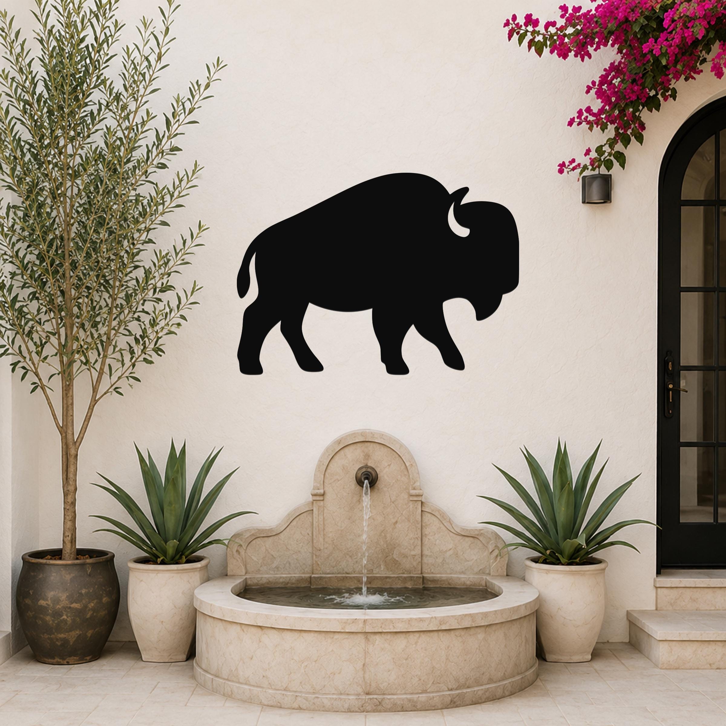 Black metal buffalo silhouette wall art mounted on a white outdoor wall above a stone fountain, flanked by potted plants and a flowering vine.