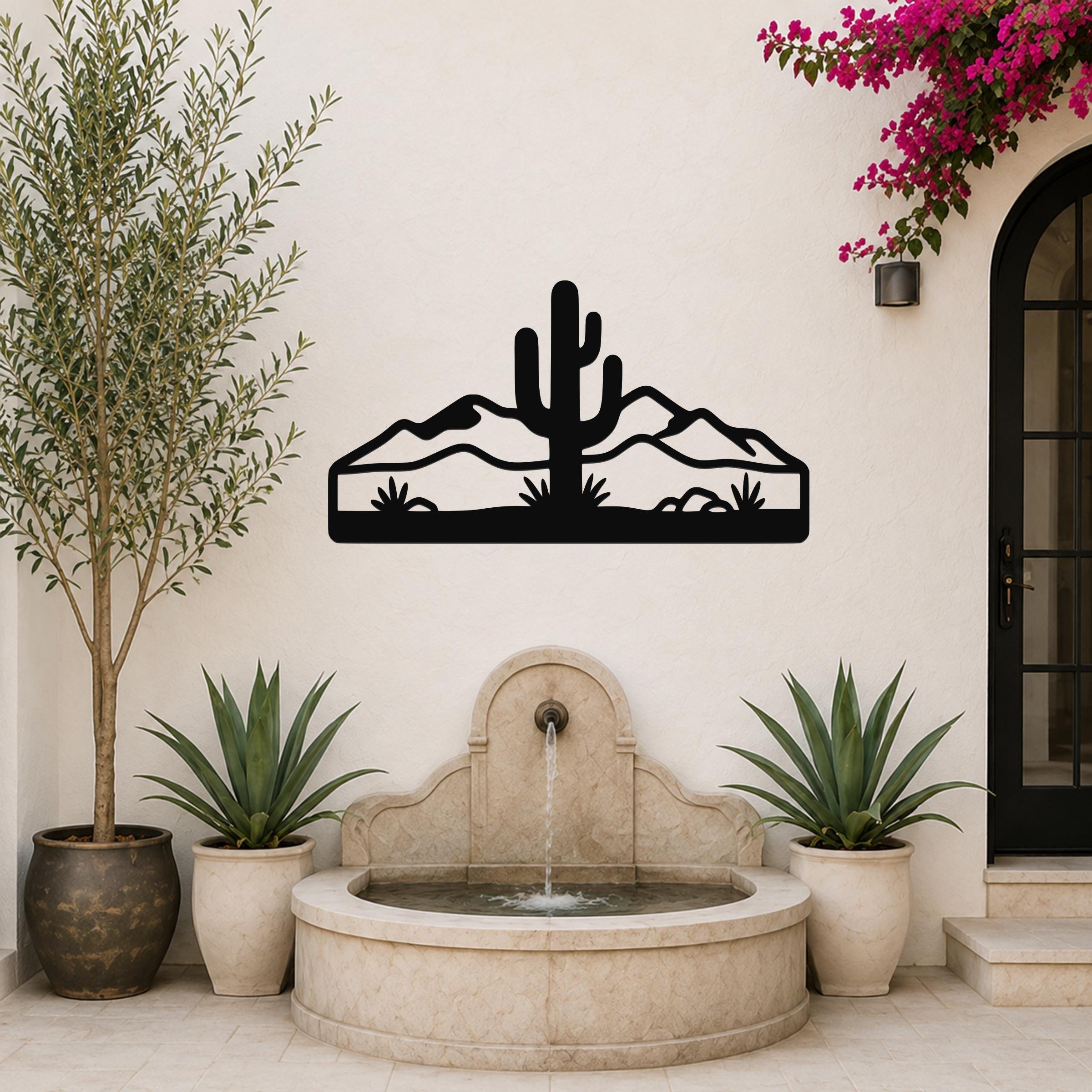 Black metal wall art featuring a saguaro cactus and mountain silhouette, mounted on a white outdoor wall above a stone fountain.