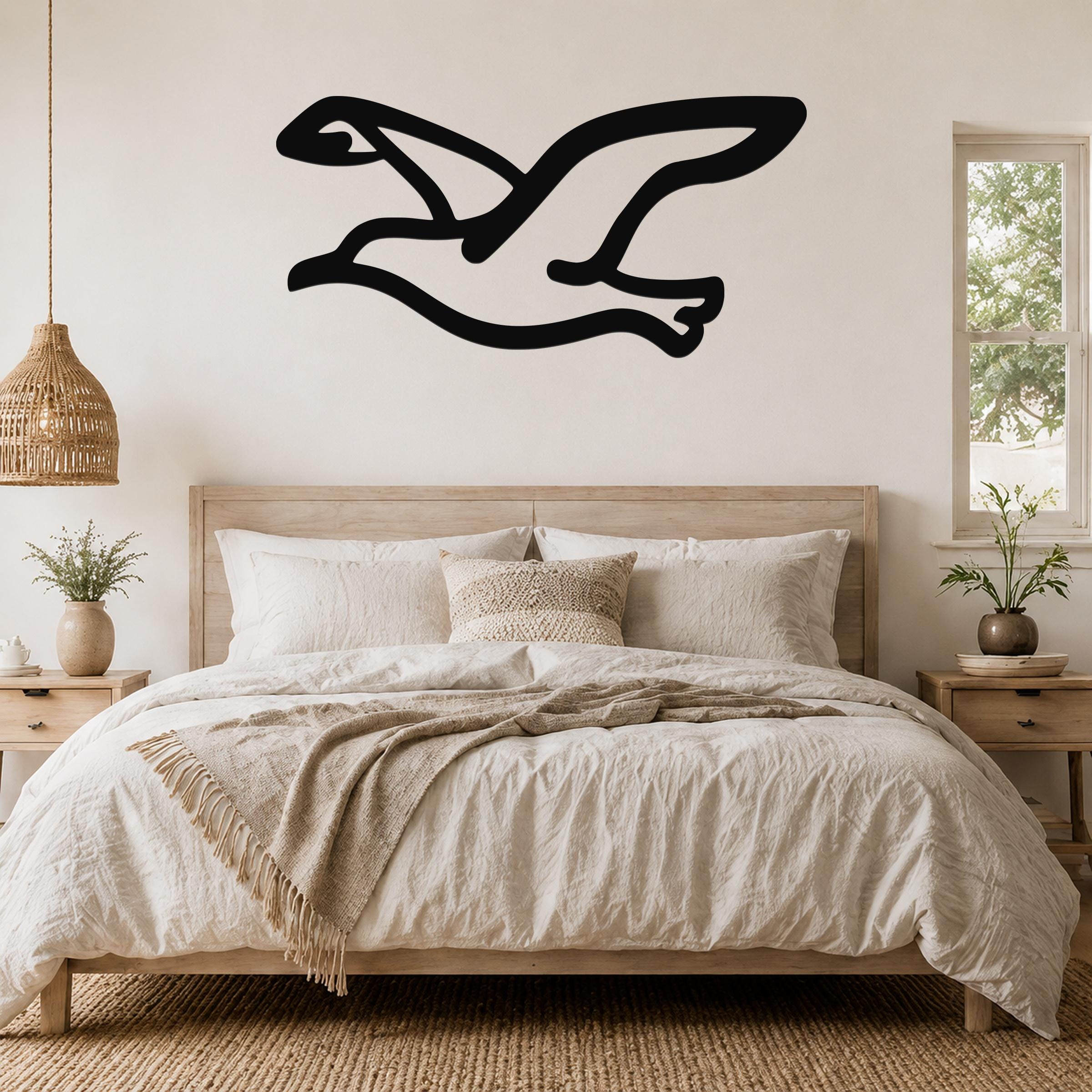 Black metal seagull wall art in a flying silhouette, mounted above a wooden bed in a neutral bedroom.