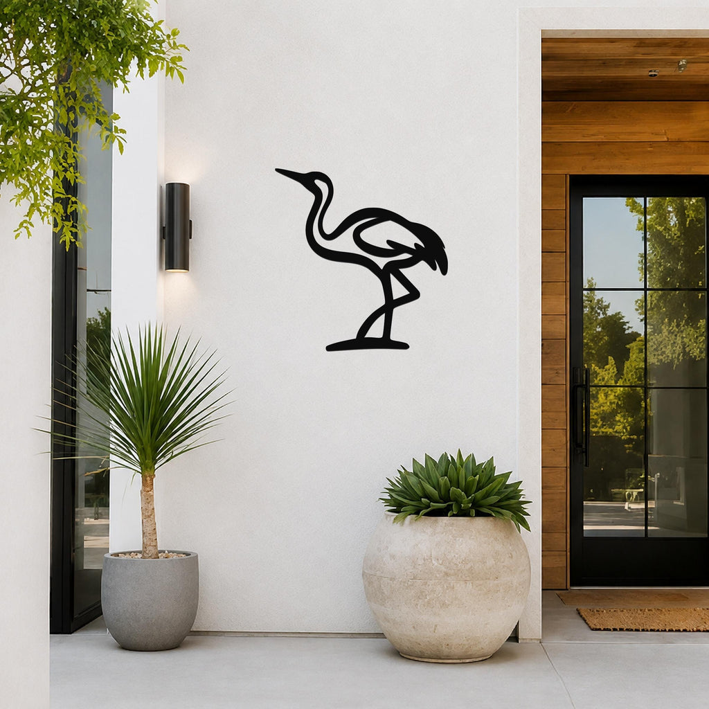 Black metal crane wall art in a minimalist silhouette style, mounted on a white exterior wall beside a glass door and potted plants.