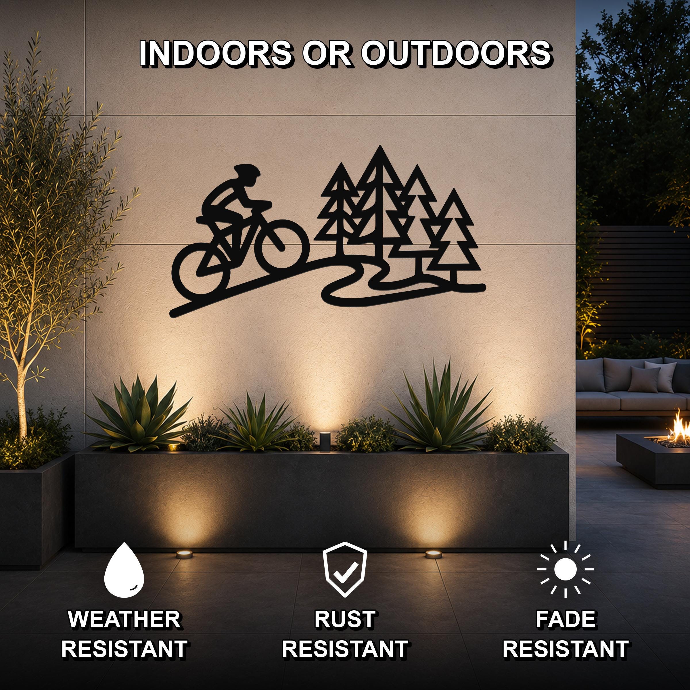 Black metal wall art featuring a cyclist silhouette and pine trees, mounted on an outdoor wall above a planter in a modern patio setting.