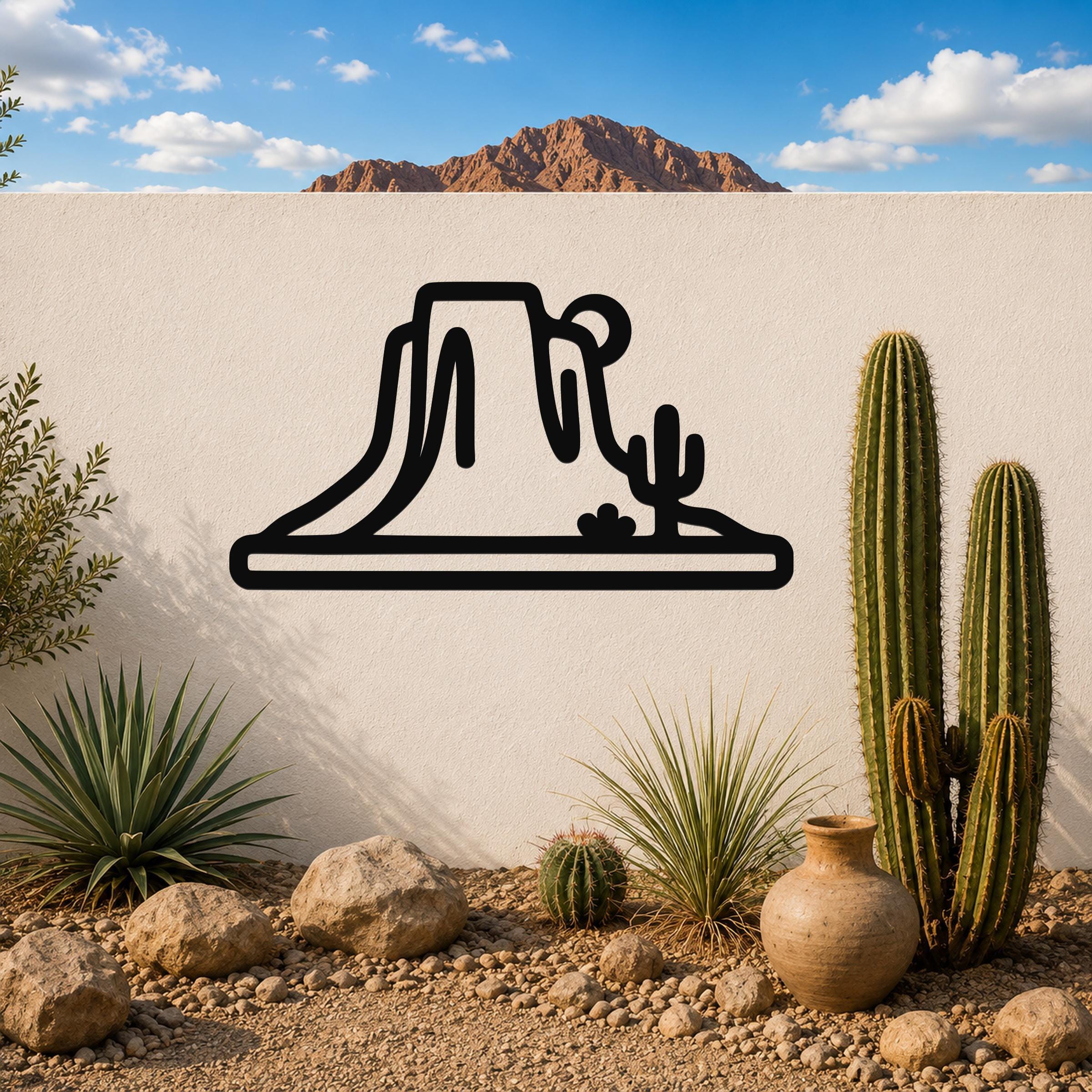 Black metal wall art silhouette of a desert mesa and cactus, mounted on a white outdoor wall above a gravel garden with real cacti and rocks.