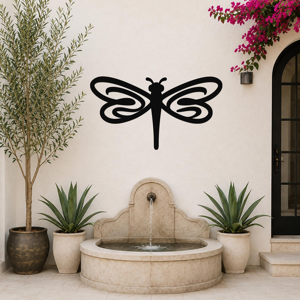 Black metal dragonfly wall art with swirling wing cutouts, mounted on a white outdoor wall above a stone fountain.