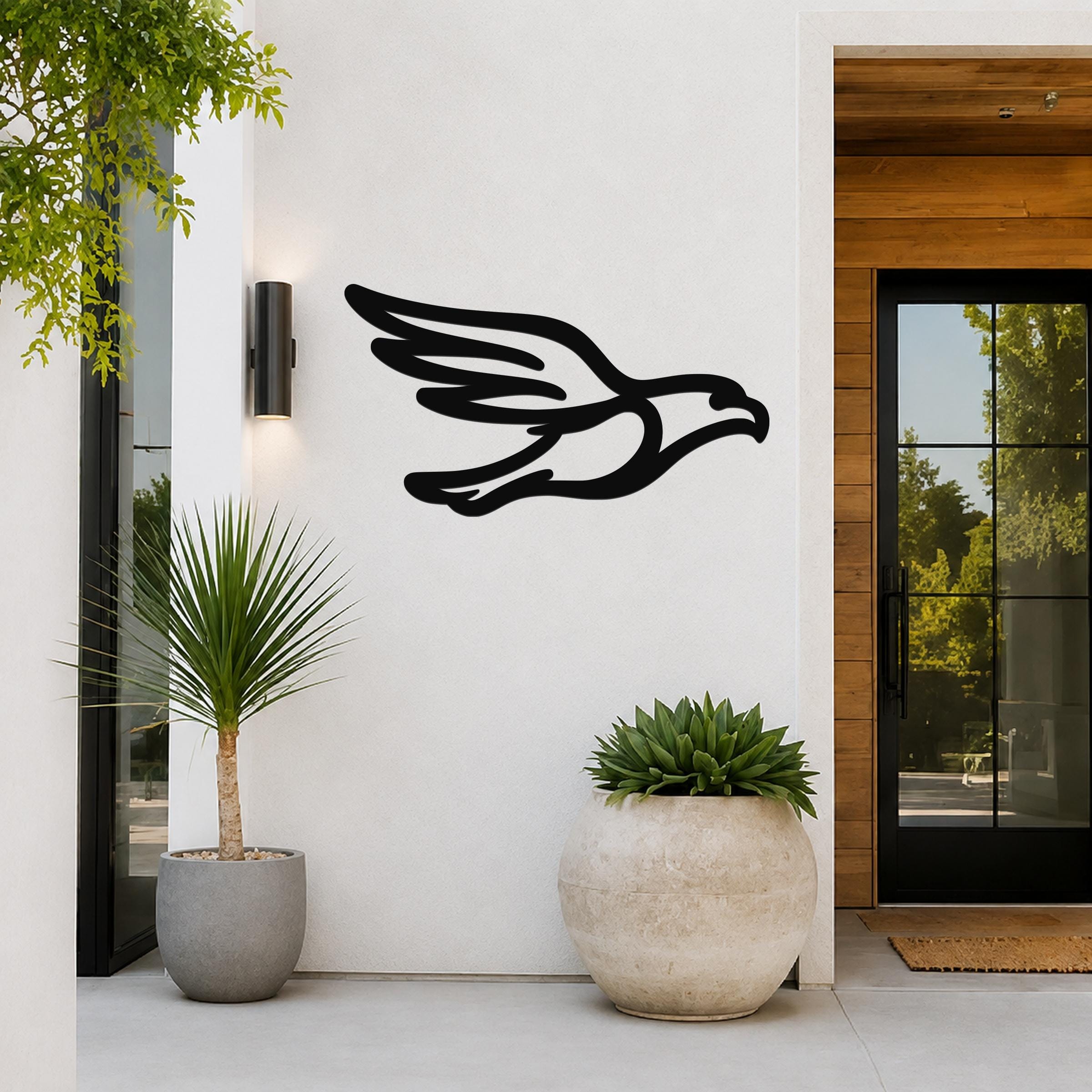 Black metal eagle wall art with a bold silhouette design, mounted on a white exterior wall beside a glass door and potted plants.