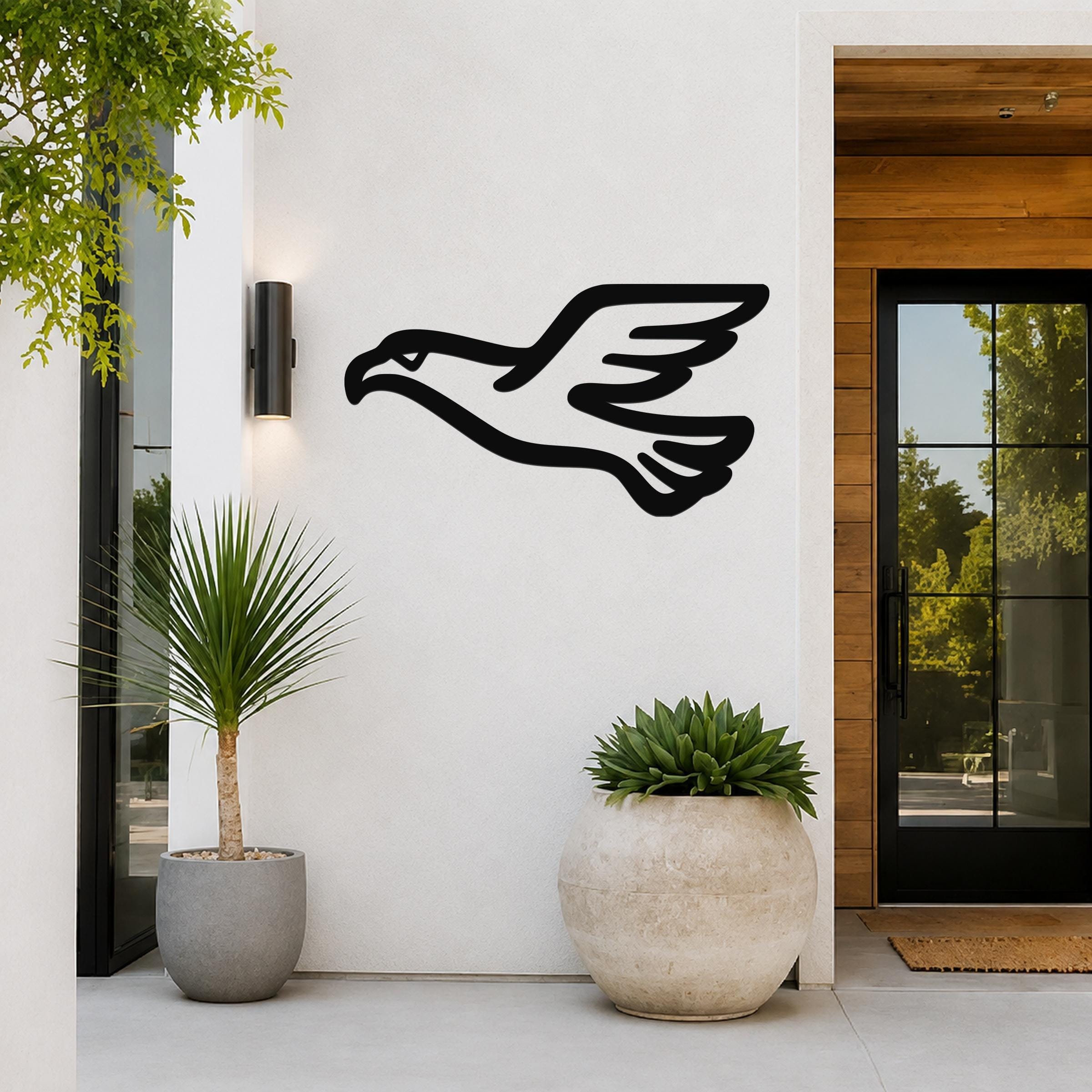Black metal eagle wall art with a soaring silhouette, mounted on a white exterior wall beside a glass door and potted plants.