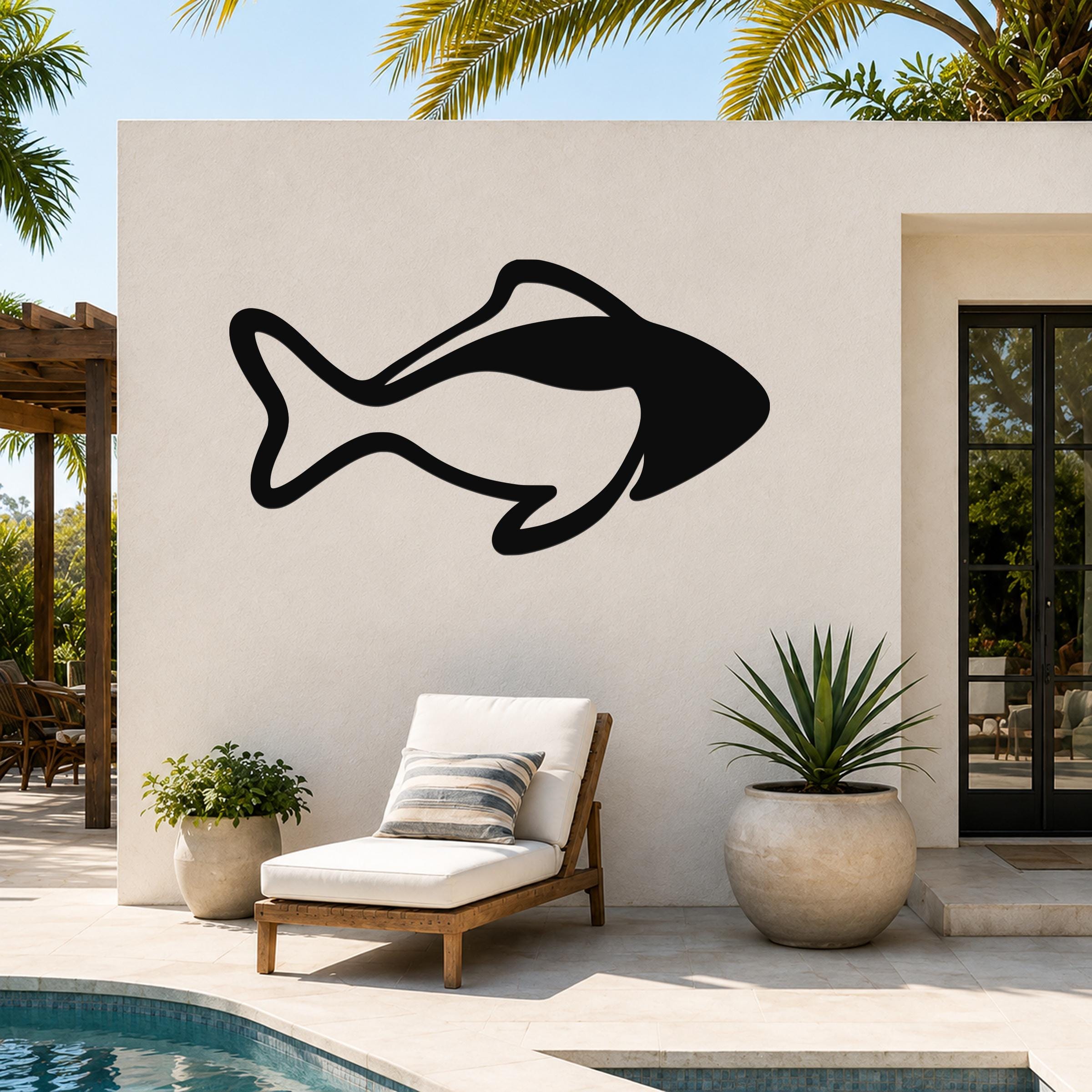 Black metal fish wall art with bold silhouette cutout, mounted on an outdoor patio wall above a lounge chair near a pool.
