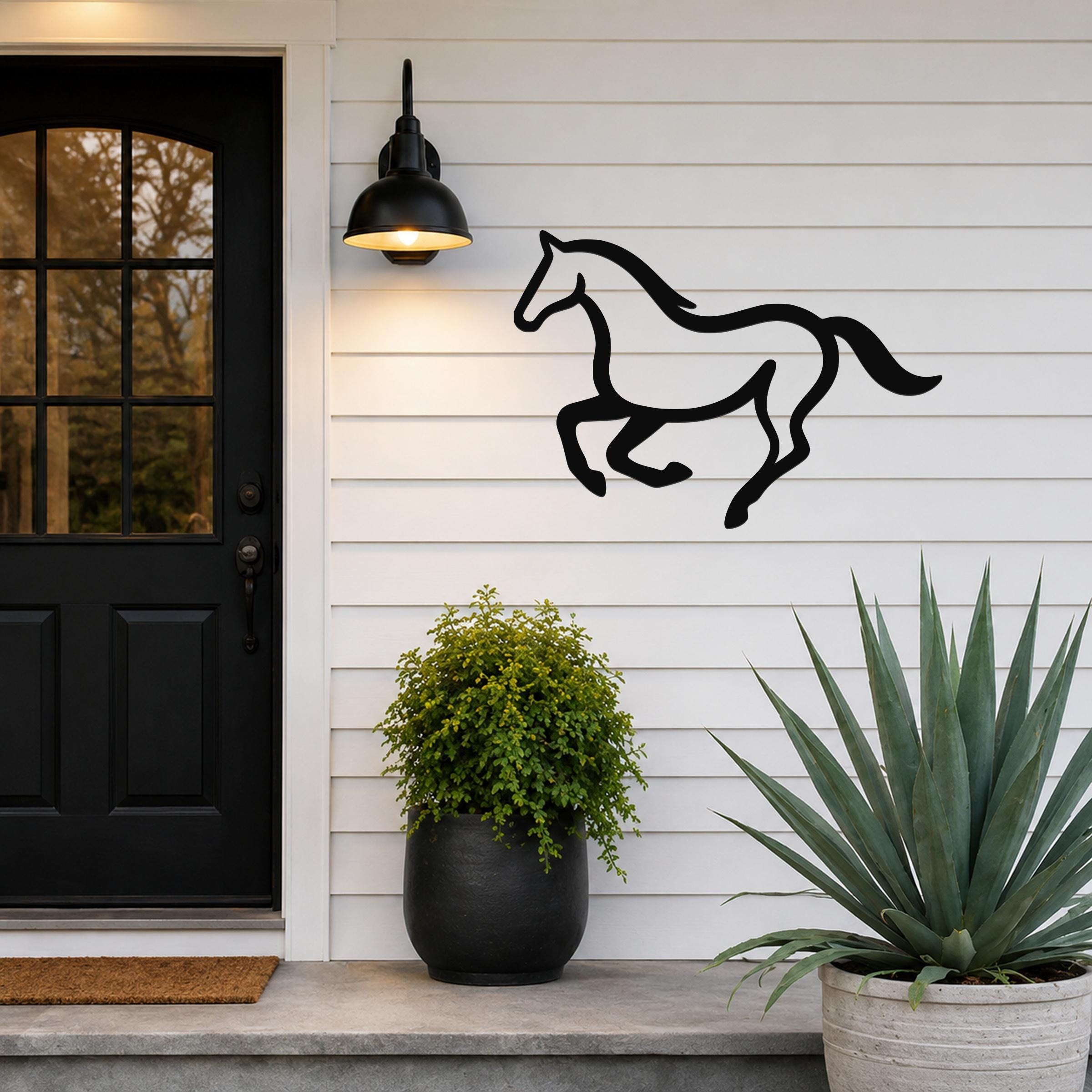 Black metal horse wall art in a running silhouette, mounted on a white siding wall beside a black front door and potted plants.
