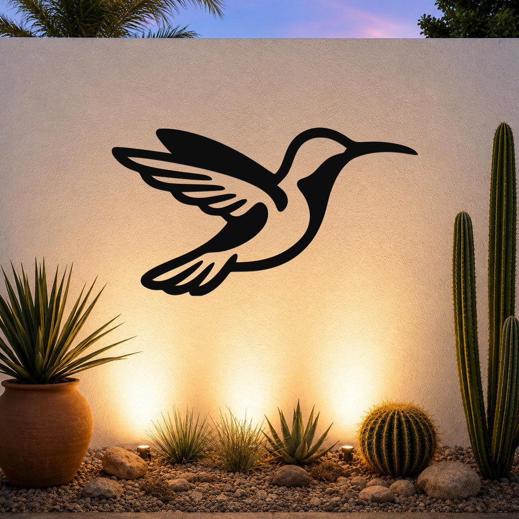 Black metal hummingbird wall art with wing and tail cutouts, mounted on an outdoor wall above a desert garden with cacti and warm uplighting.
