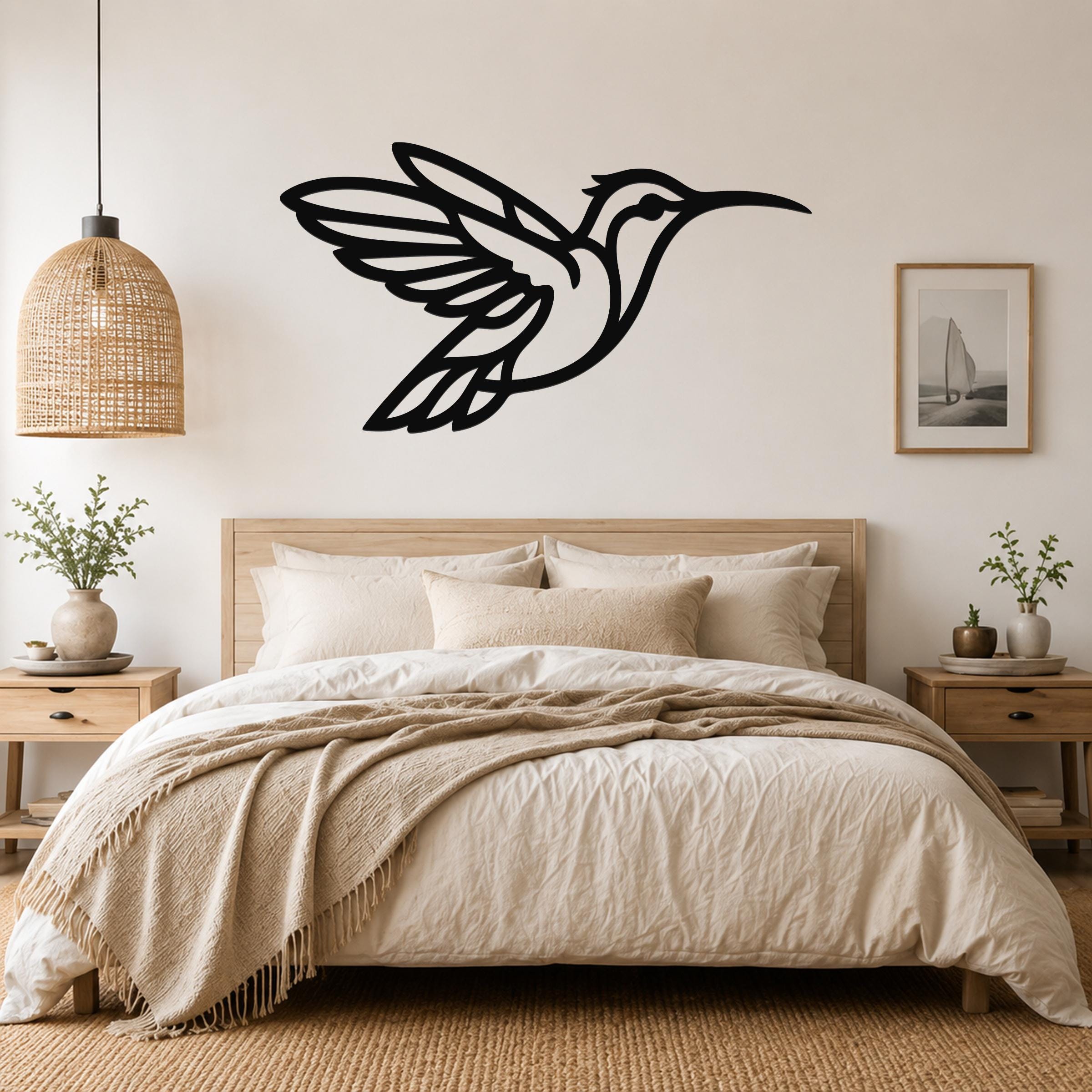 Black metal hummingbird wall art with wing and body cutouts, mounted above a bed in a neutral bedroom.