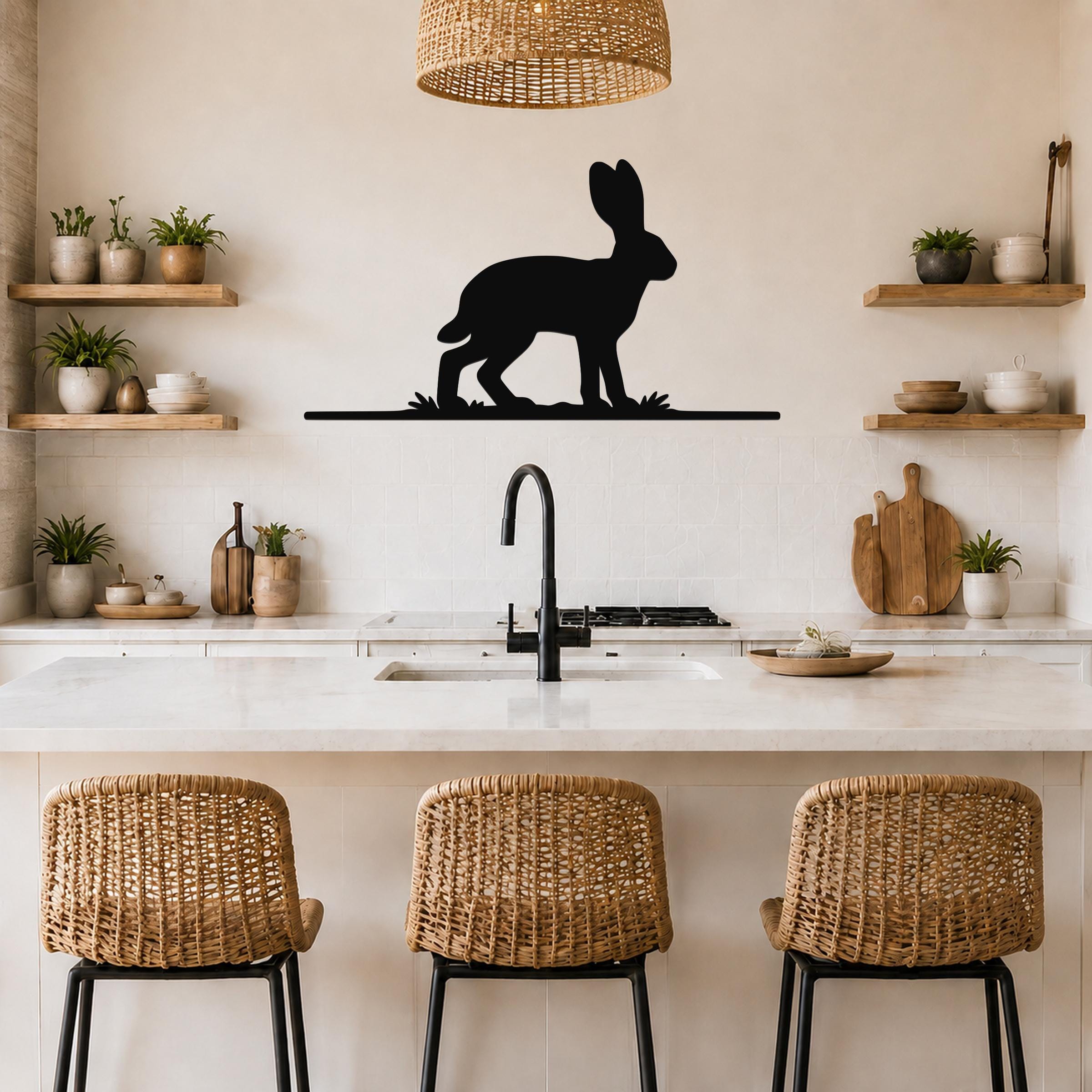 Black metal jackrabbit wall art silhouette mounted above a kitchen counter, featuring a standing hare with grass cutouts, in a modern rustic kitchen with woven chairs and open shelves.