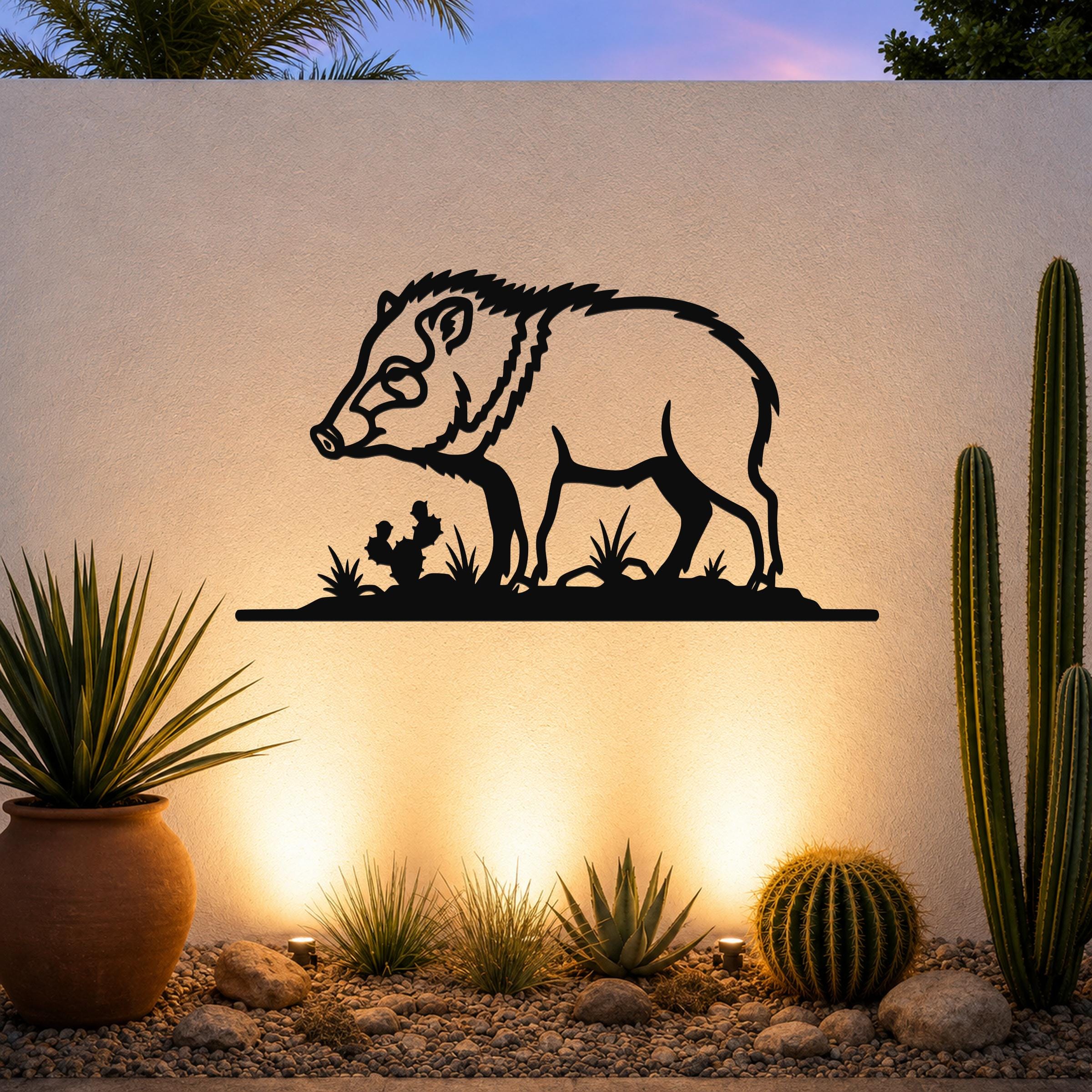 Black metal javelina wall art with a silhouette design of the animal and desert plants, mounted on an outdoor wall above a gravel garden with cacti and warm uplighting.