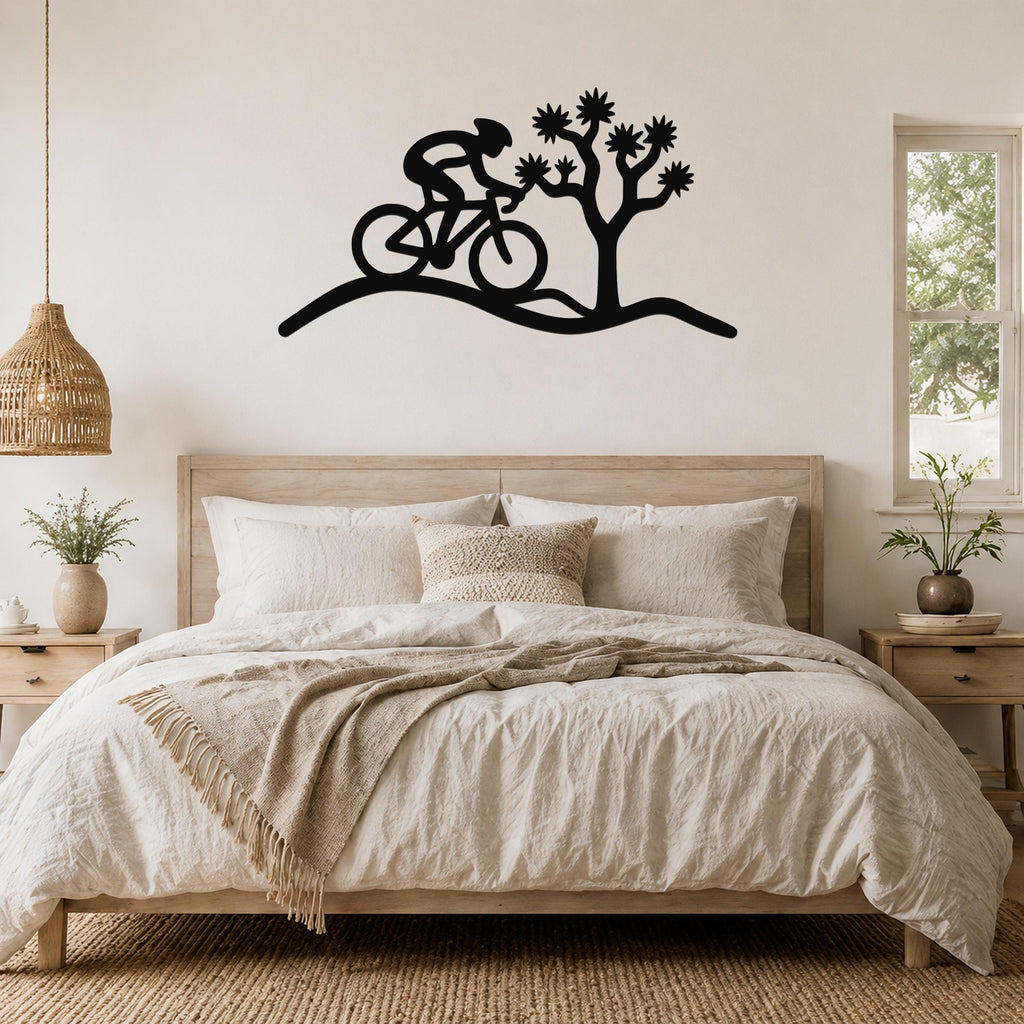 Black metal wall art silhouette of a cyclist riding past a Joshua tree, mounted above a bed in a bedroom.