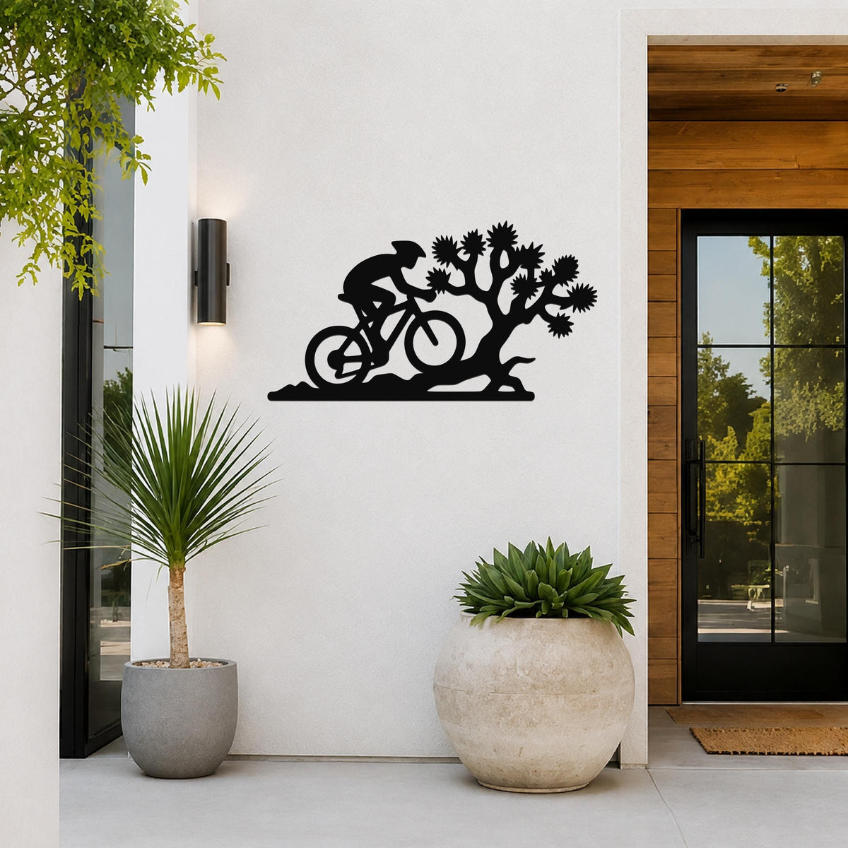 Black metal wall art silhouette of a cyclist riding past a Joshua tree, mounted on a white exterior wall beside a glass door and potted plants.