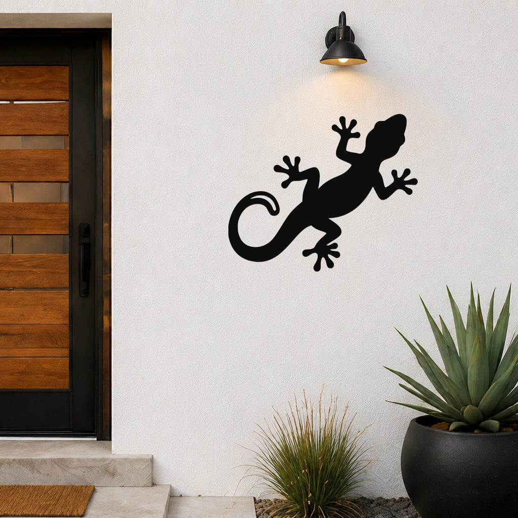 Black metal gecko wall art silhouette mounted on a white exterior wall above a potted succulent, illuminated by a black outdoor sconce light.