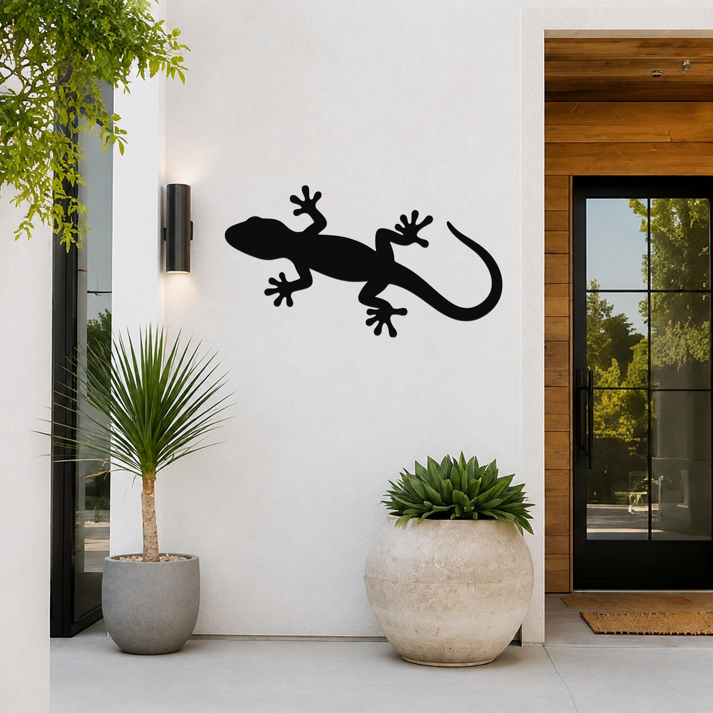 Black metal gecko wall art silhouette mounted on a white exterior wall beside a glass door and potted plants.